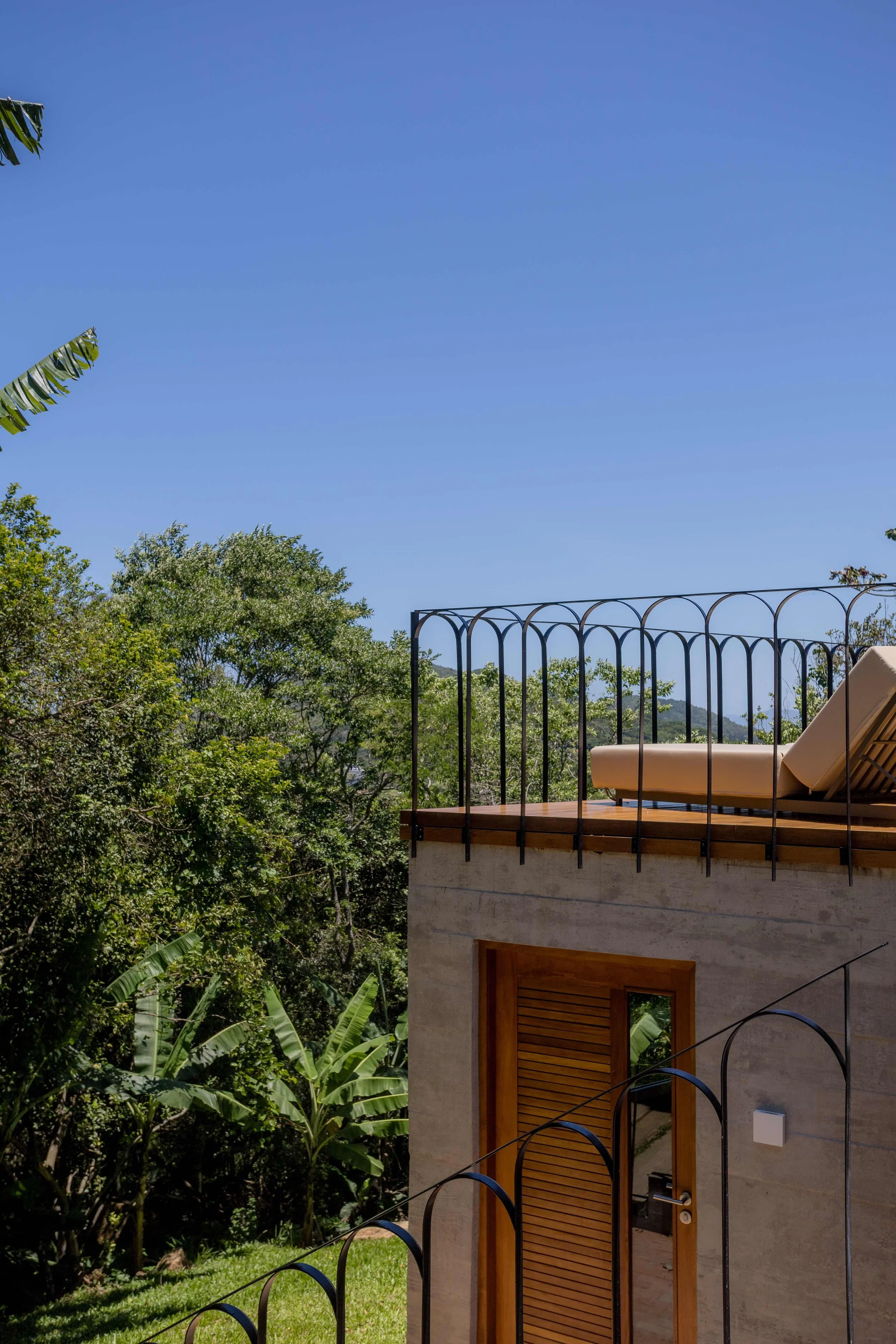 A house with a rooftop patio containing a lounge chair, surrounded by green trees and set against a clear blue sky.