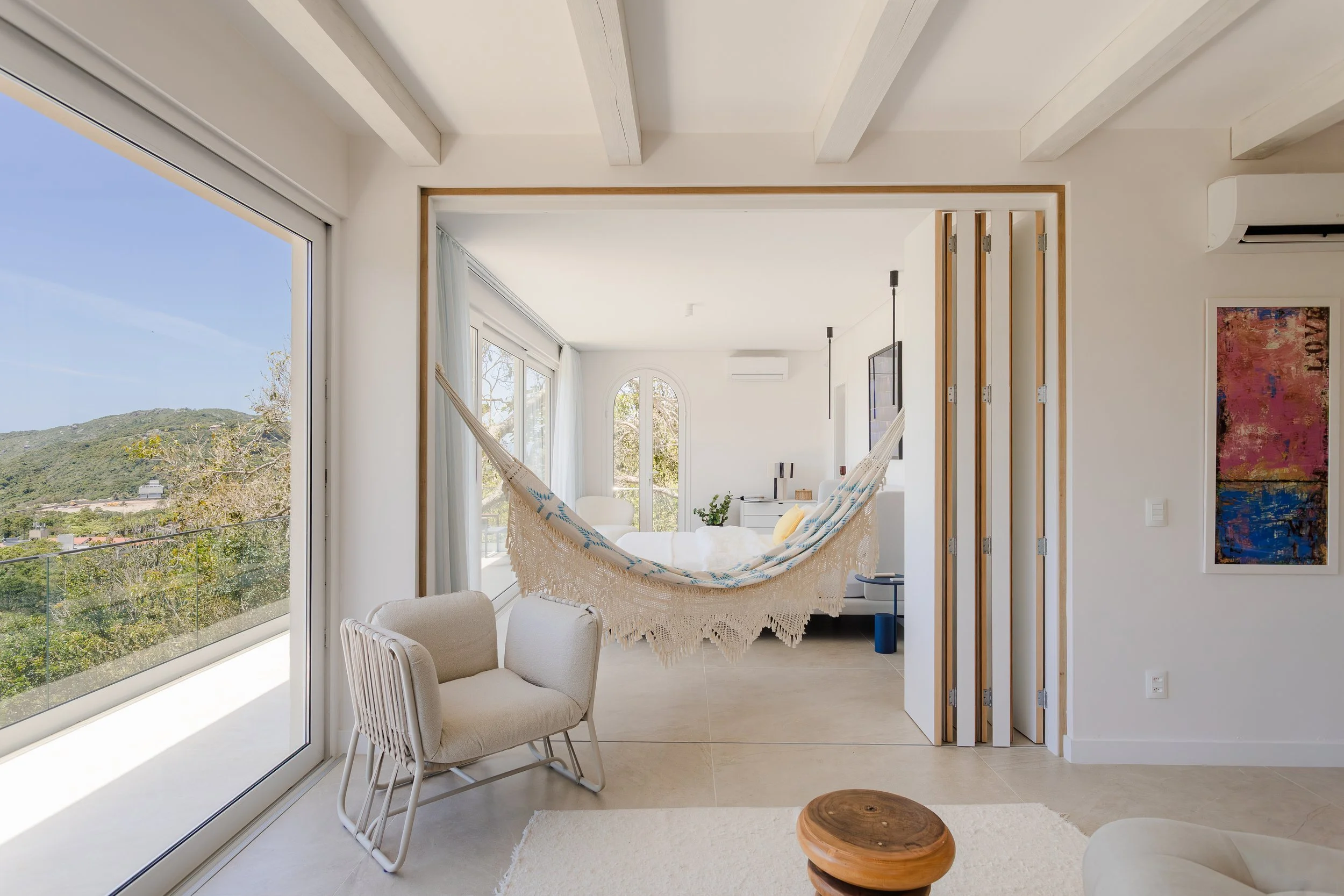 Bright living room with a hammock hanging in between two walls, large windows with white curtains, contemporary furniture, and a scenic view of hills and trees outside.