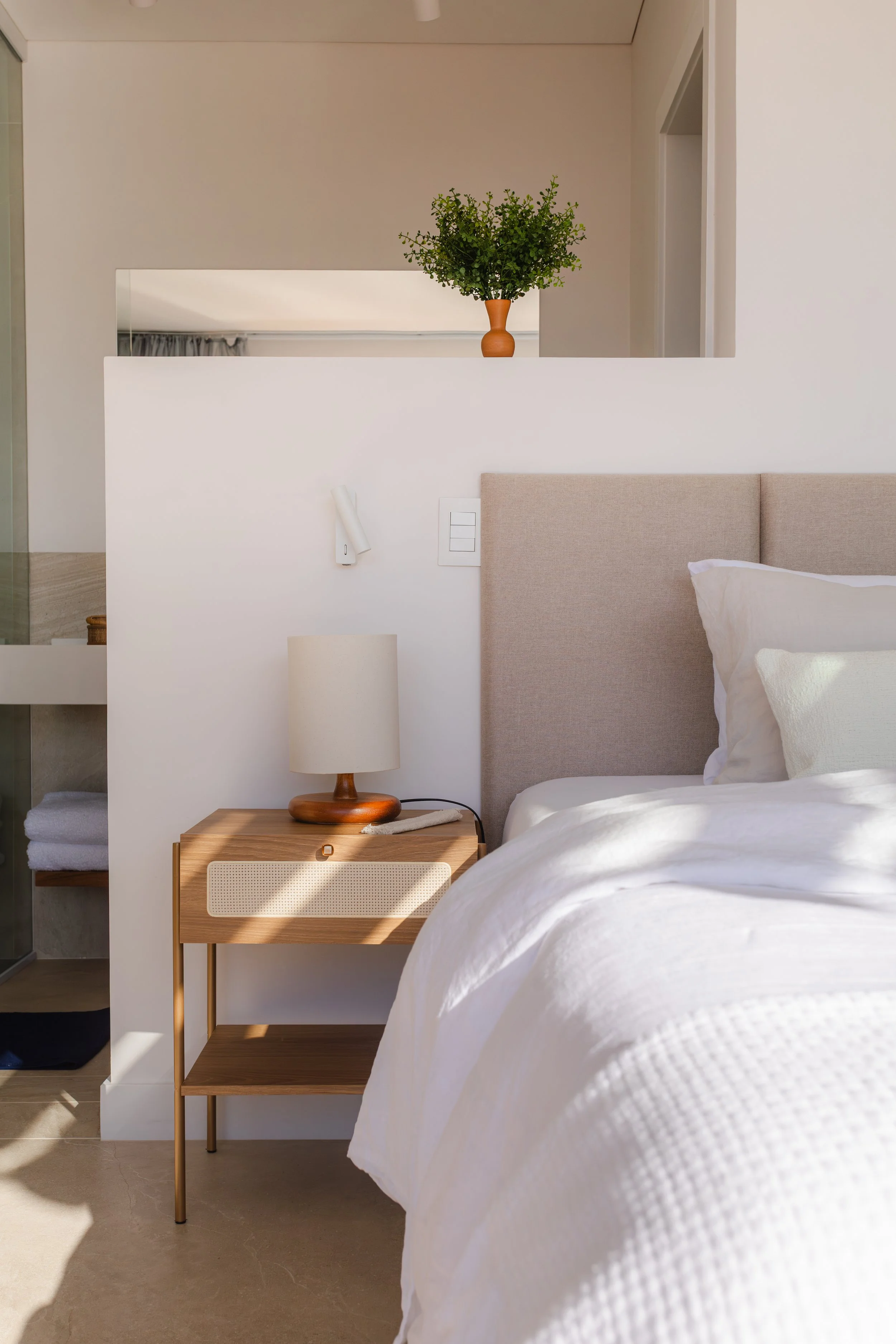 A modern bedroom with a beige upholstered headboard, white bedding, and a wooden nightstand with a lamp. Behind the bed, there's a half wall with a decorative vase and greenery on top, and part of an open bathroom with towels.