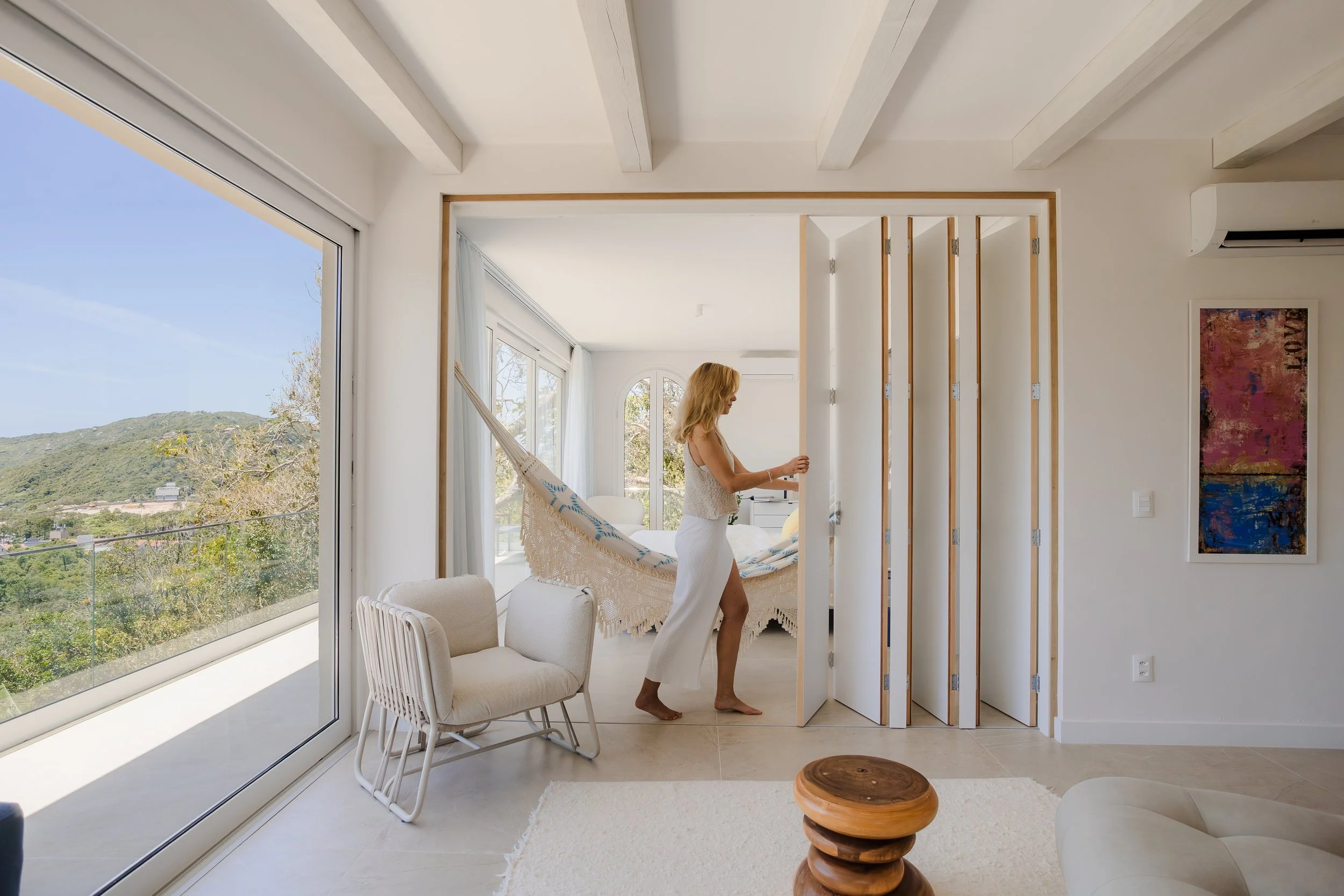 A woman standing in a bright, modern bedroom, opening a sliding door to a balcony with a view of trees and hills, with natural light filling the room.