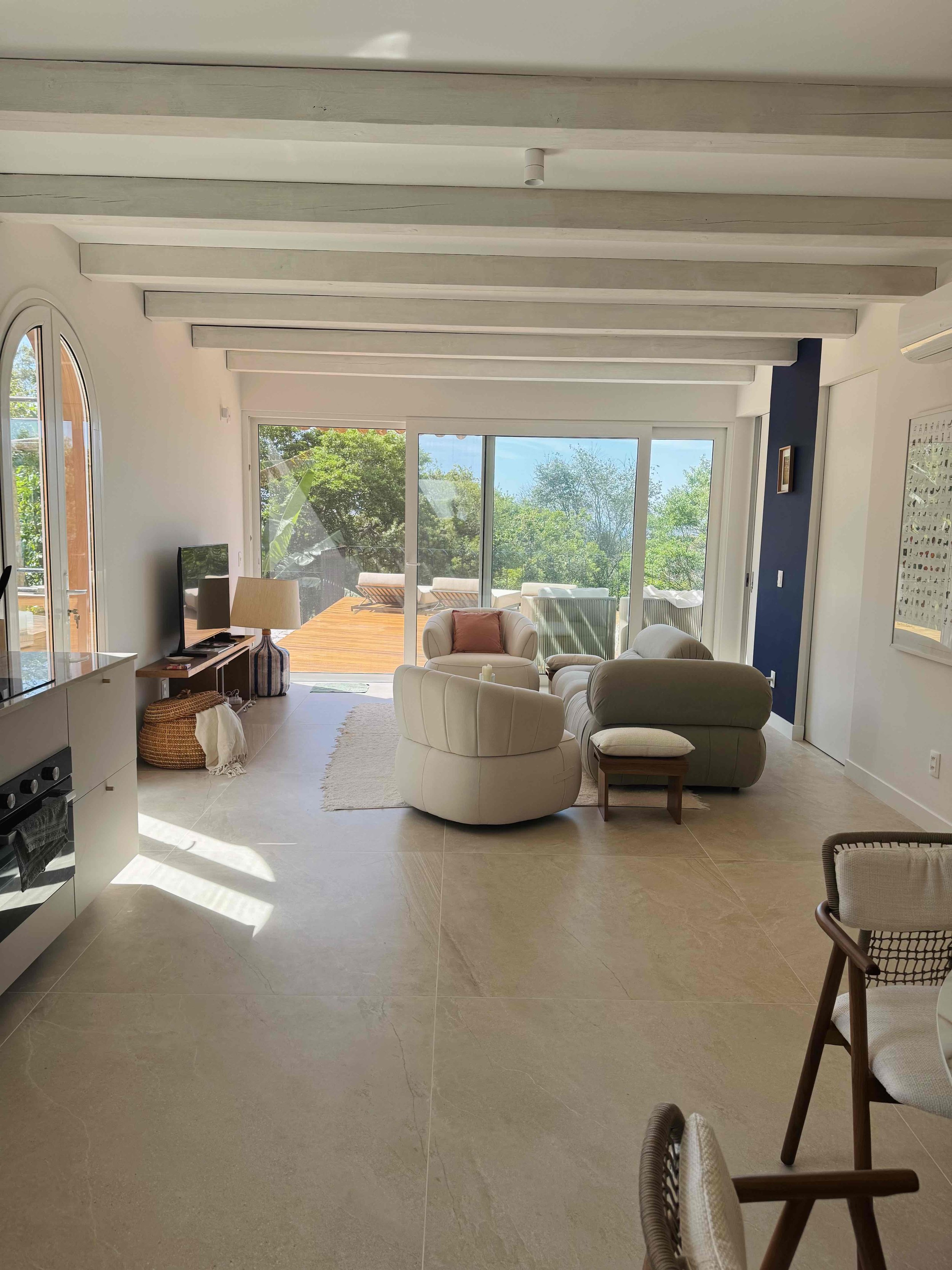 Bright living room with beige furniture, large sliding glass door leading to outdoor deck with greenery, modern decor, and a partial view of the kitchen.