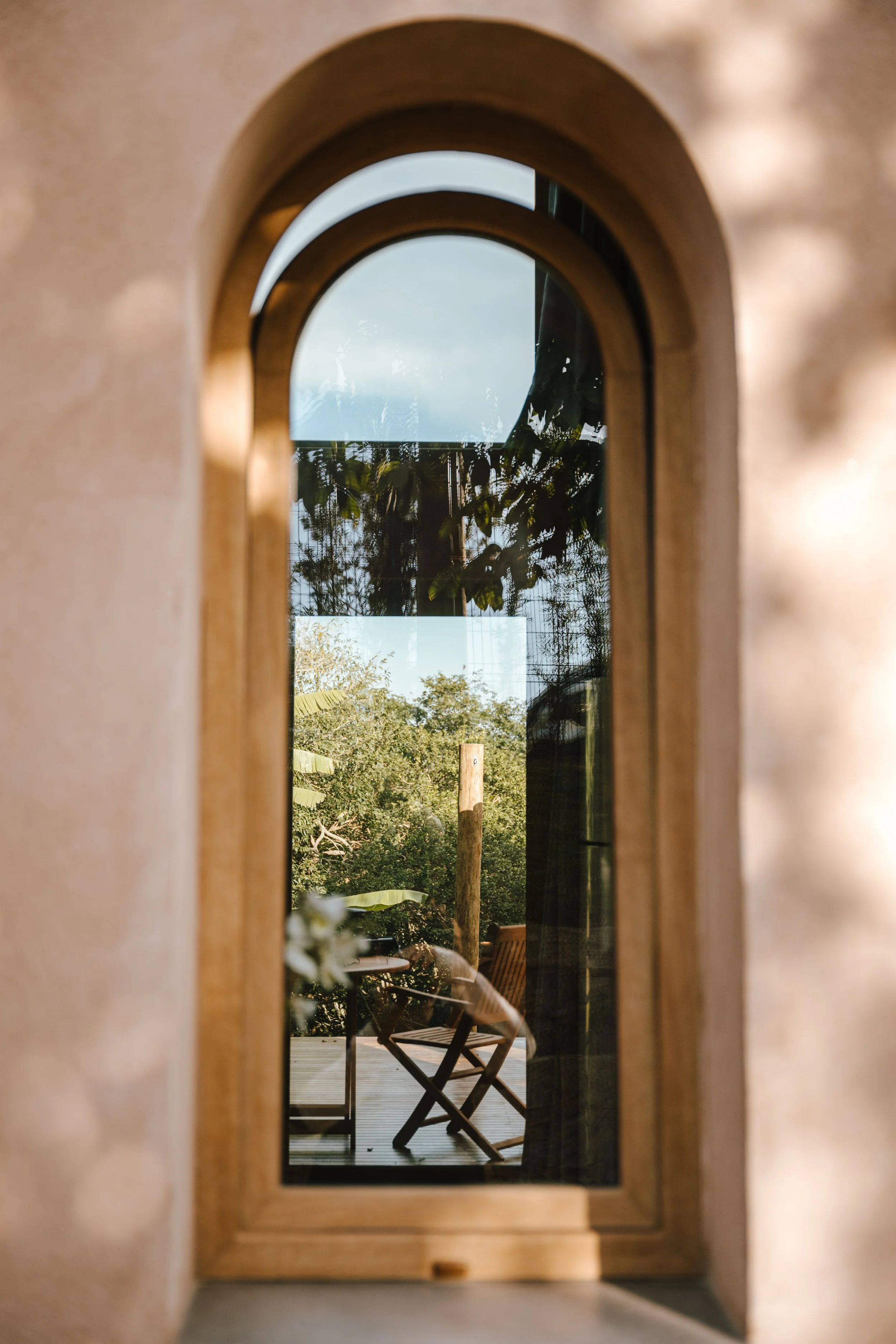 A view through a narrow arched outdoor window showing a wooden deck with a chair and table, surrounded by lush greenery and a clear sky.