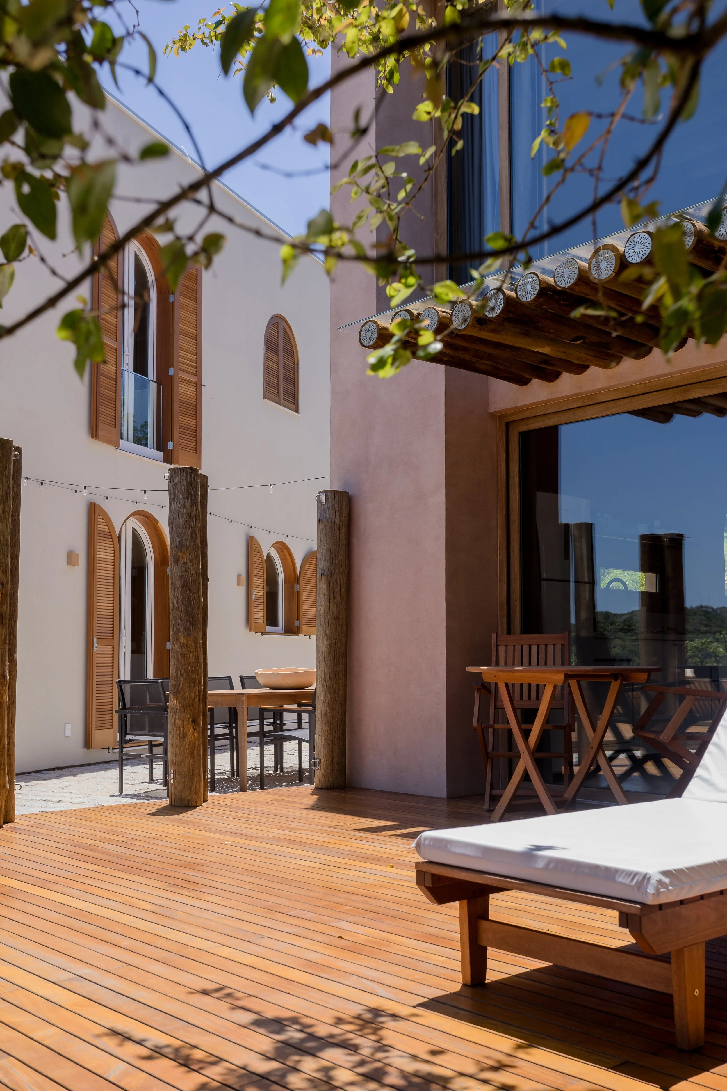 View of a sunny wooden patio with a white lounge chair, a small wooden table with chairs outside a modern building with arched windows and wooden shutters, climbing plants, and string lights.