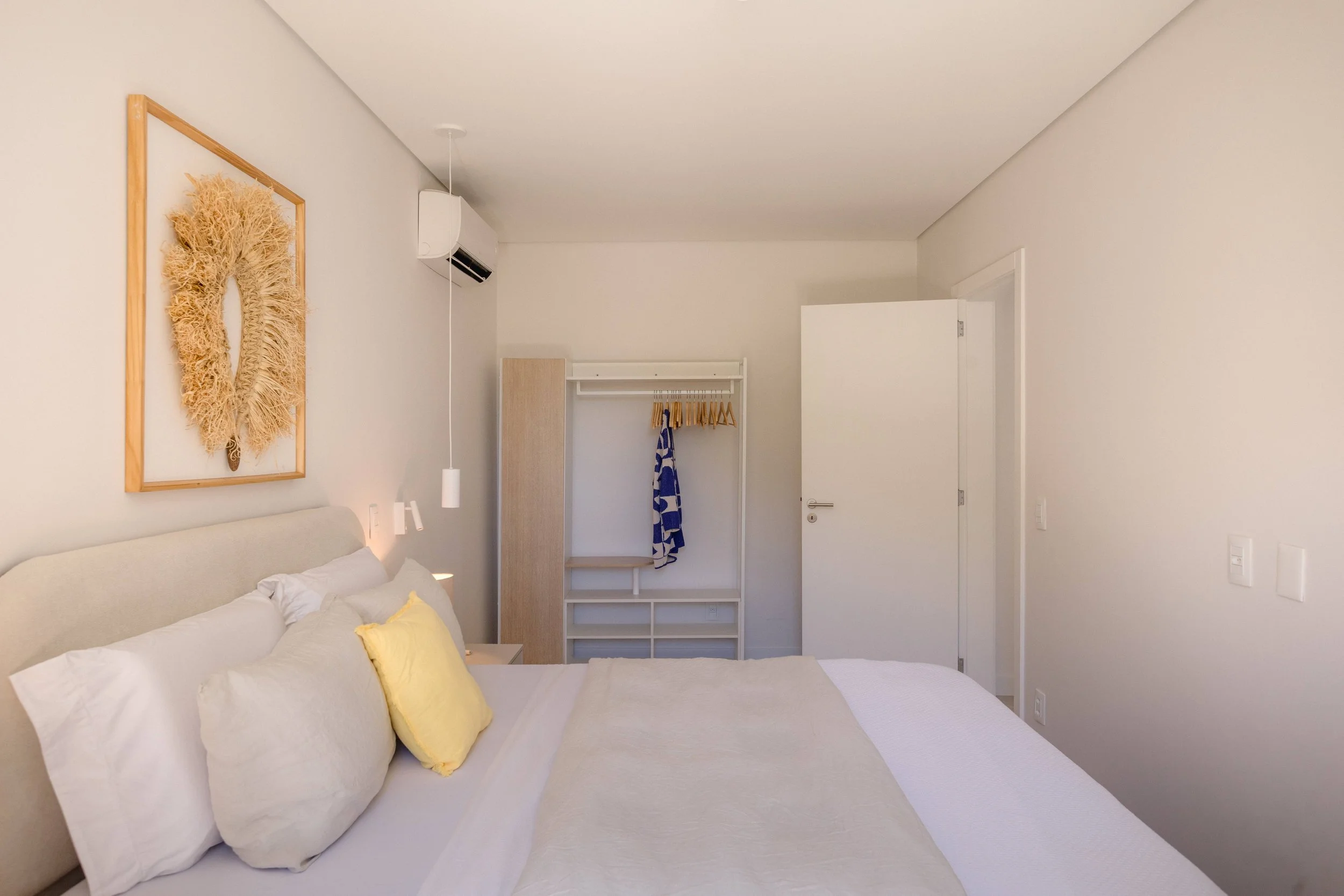 A minimalistic bedroom with a large bed, decorative pillows, and a bedside table. There is a wall art piece above the headboard, an open closet with hanging clothes, and a door on the right side. The room is decorated in neutral tones.