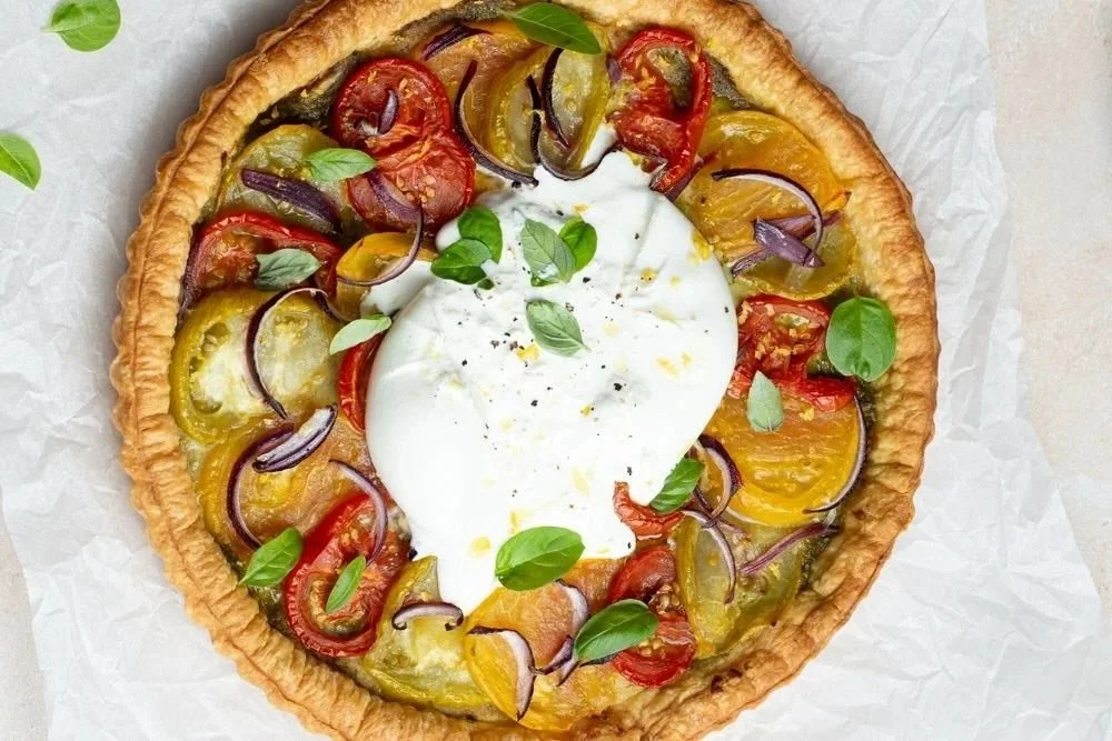 Vegetable tart with a puff pastry dough , topped with cherry tomatoes, yellow and red bell peppers, red onion slices, fresh basil leaves, and a creamy burrata in the center.