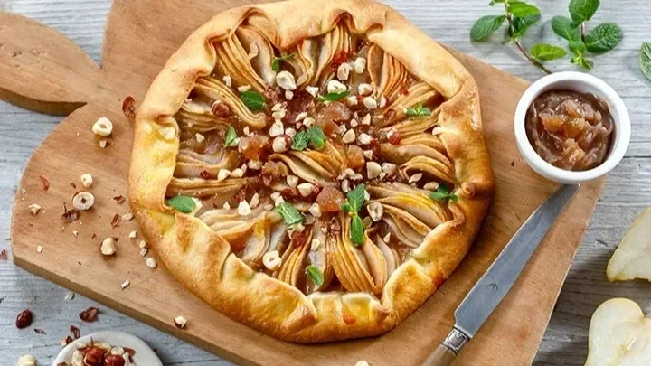 Rustic apple galette with caramelized apple slices, chopped nuts, and fresh mint on a wooden board, with a small bowl of caramelized fruit topping.