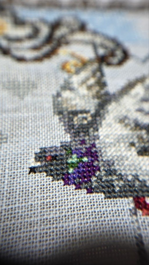 Close-up of a cross-stitch embroidery showing a design with black, purple, green, and red threads on white fabric.