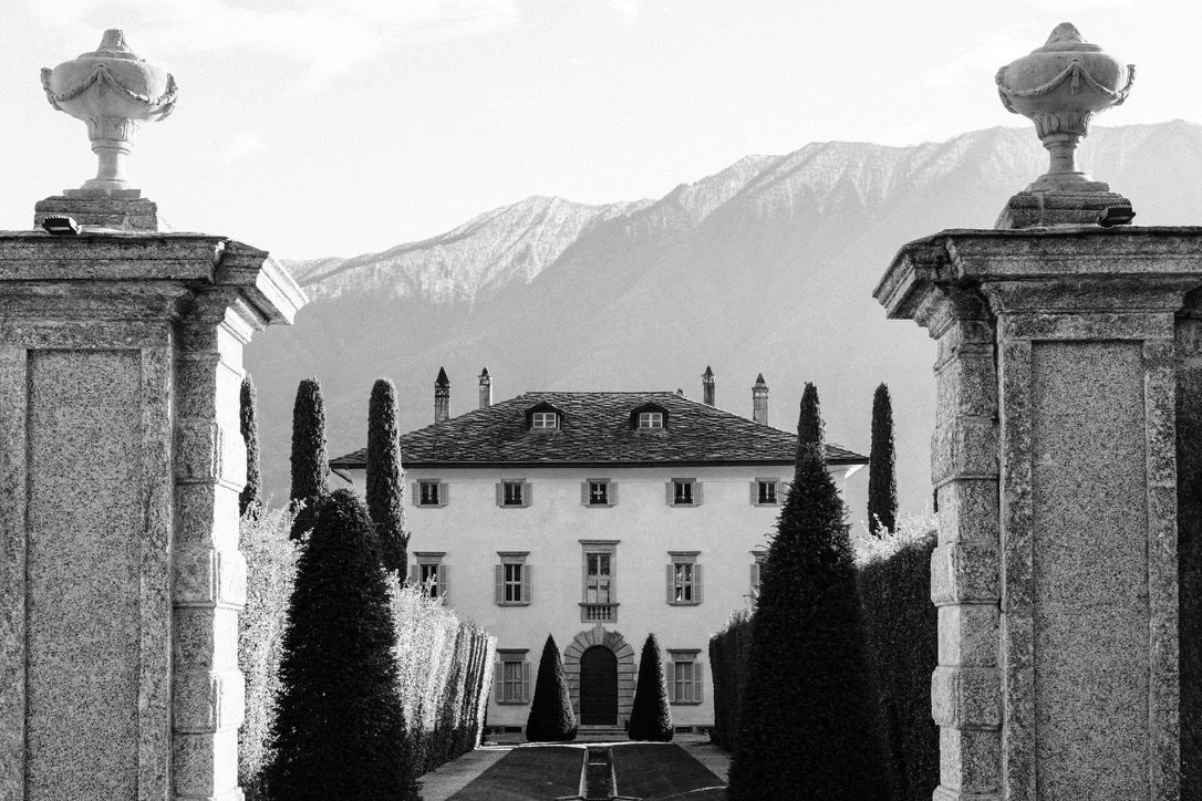 A grand mansion with a dark roof, surrounded by tall, narrow cypress trees, and flanked by stone pillars with large urns on top, with mountain peaks in the background.