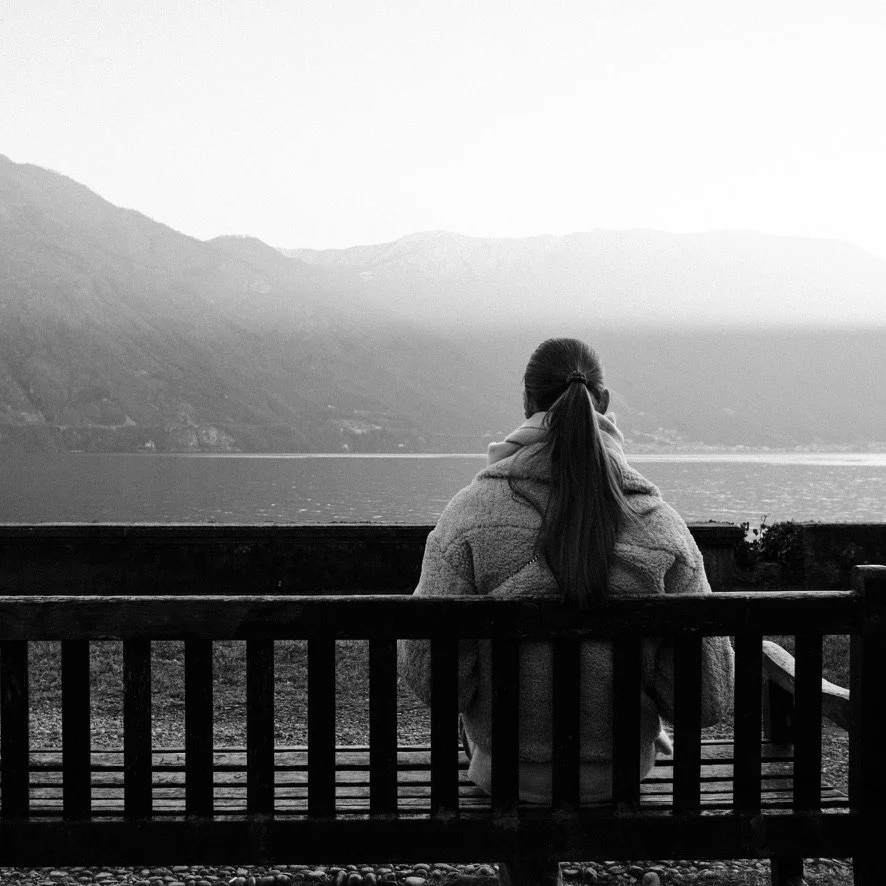 A woman with long hair in a ponytail, wearing a coat, sitting alone on a bench, overlooking a lake surrounded by mountains in black and white.