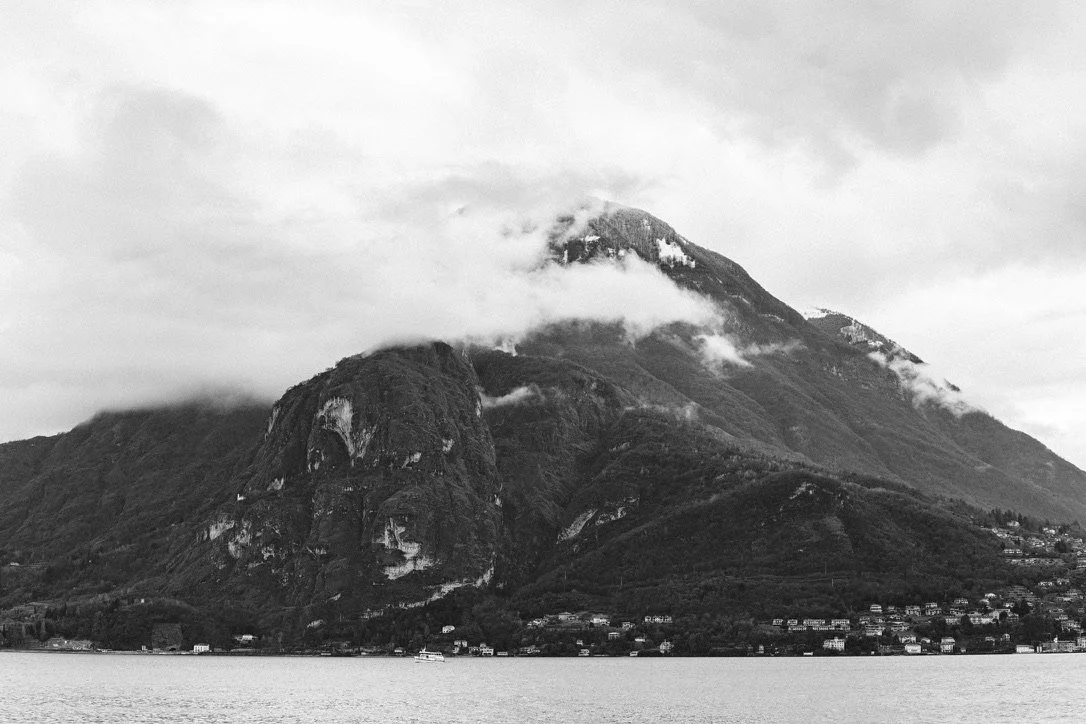 A black and white photograph of a large mountain with clouds near its peak, overlooking a body of water with small houses at the base.
