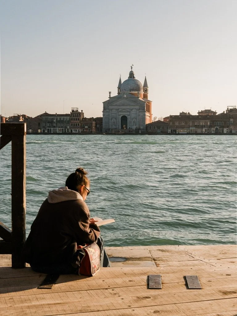 A person sitting by the water on a wooden dock, reading a book, with a historic building and a row of houses across the water, in Venice, Italy.