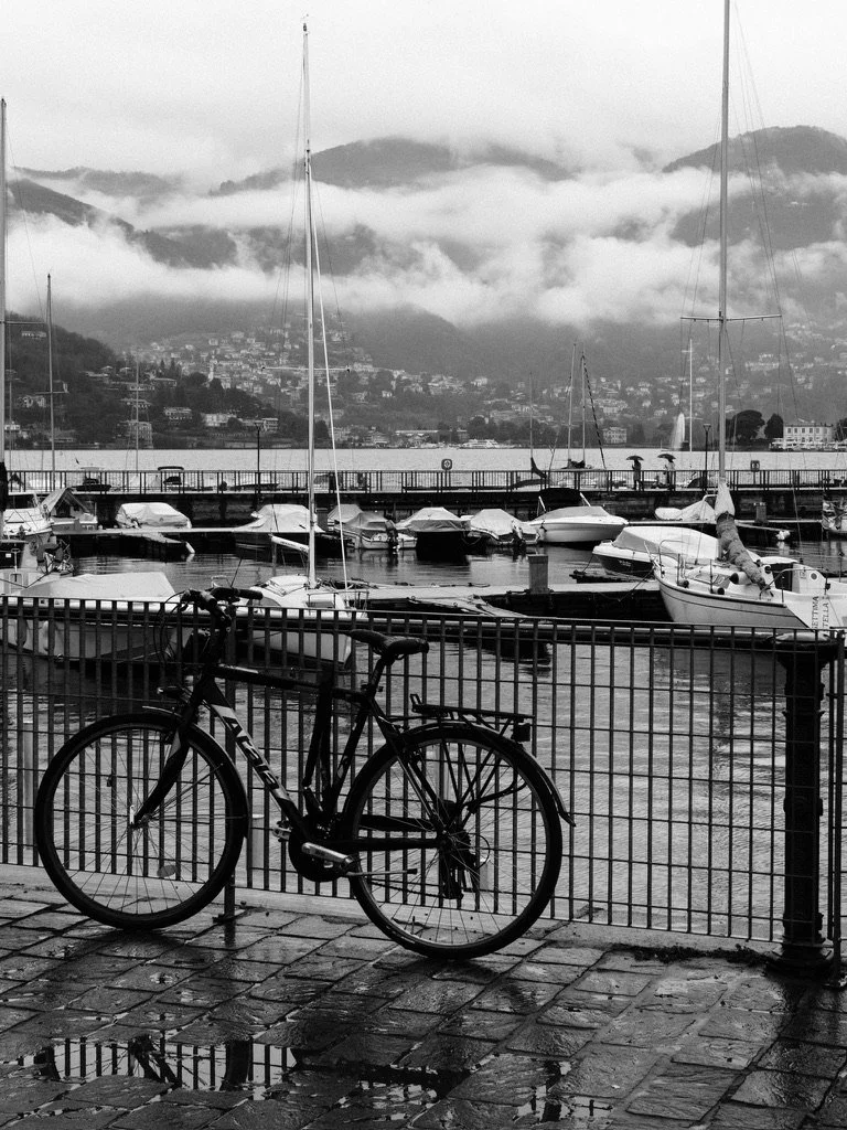 Black and white photo of a bicycle leaning against a metal fence by a marina, with docked sailboats and a cloudy mountain landscape in the background.