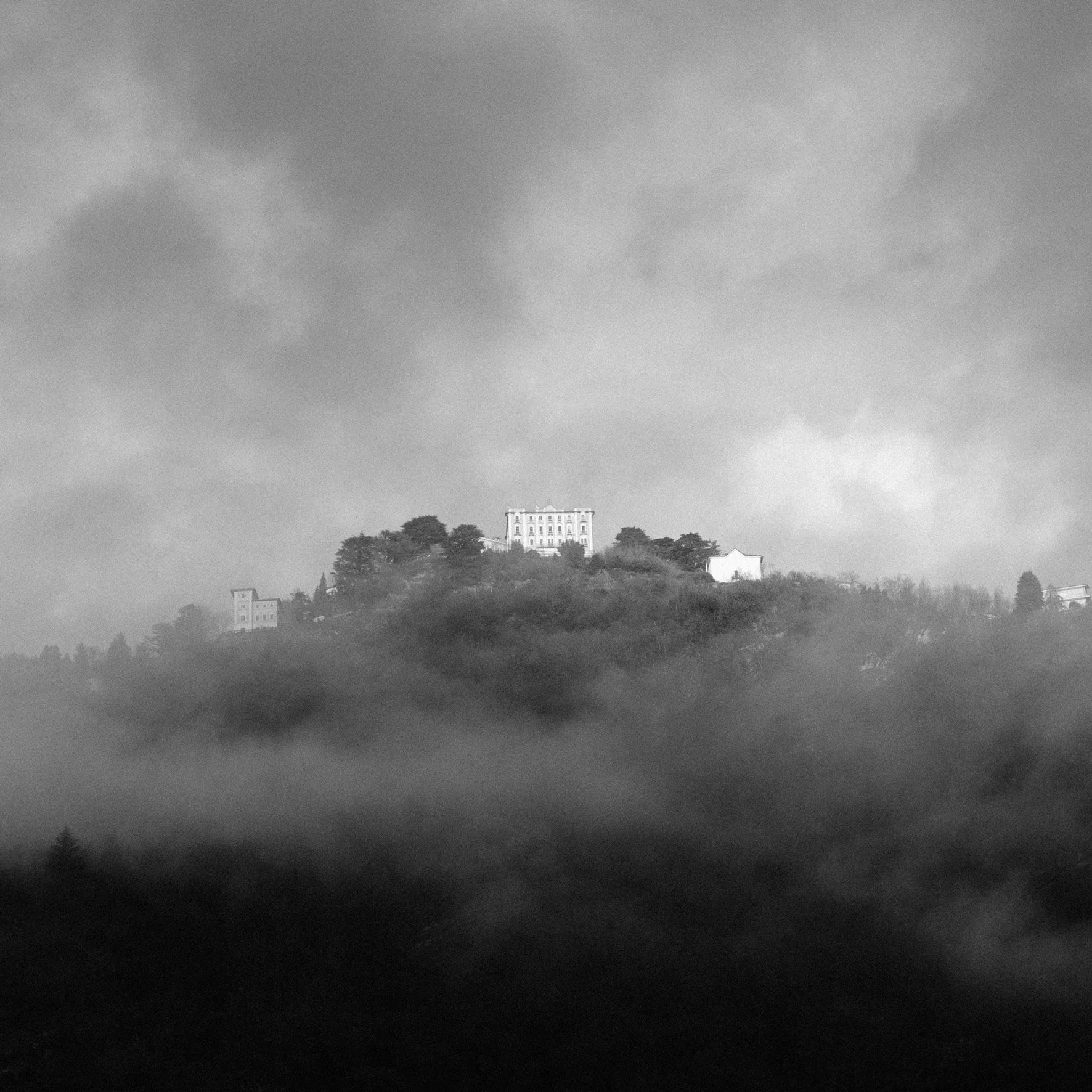 A black and white photograph of a hilltop with structures and trees, shrouded in fog.