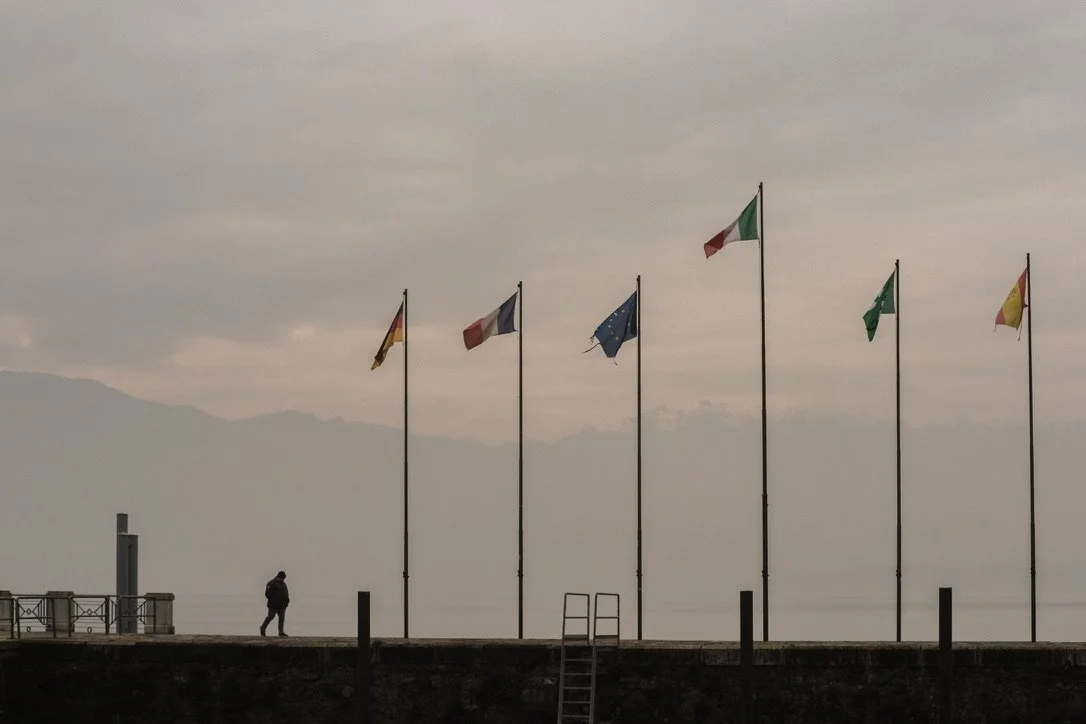 A person walking along a seaside promenade with six flags on tall poles, including flags of Germany, France, the European Union, Italy, Saudi Arabia, and Spain, during cloudy weather.