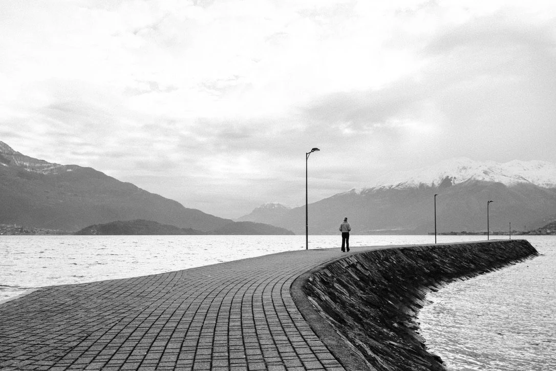A person standing on a curved pier by a body of water, with mountains in the background, overcast sky.