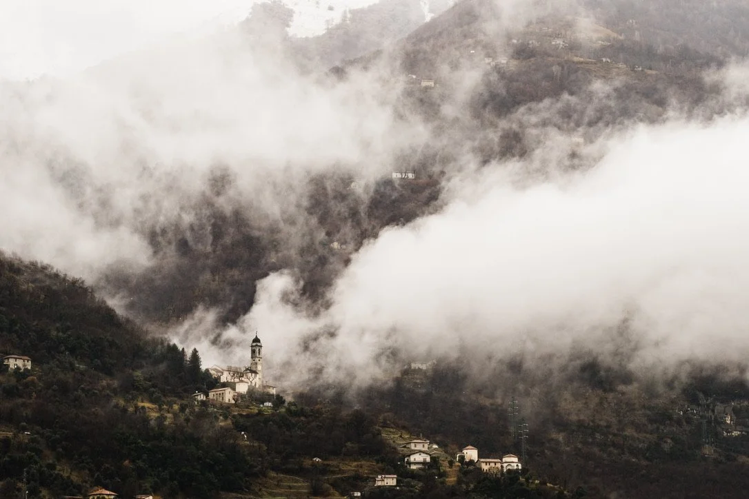 Mountain landscape with scattered houses and a church steeple in the foreground, partially obscured by thick fog or low clouds.