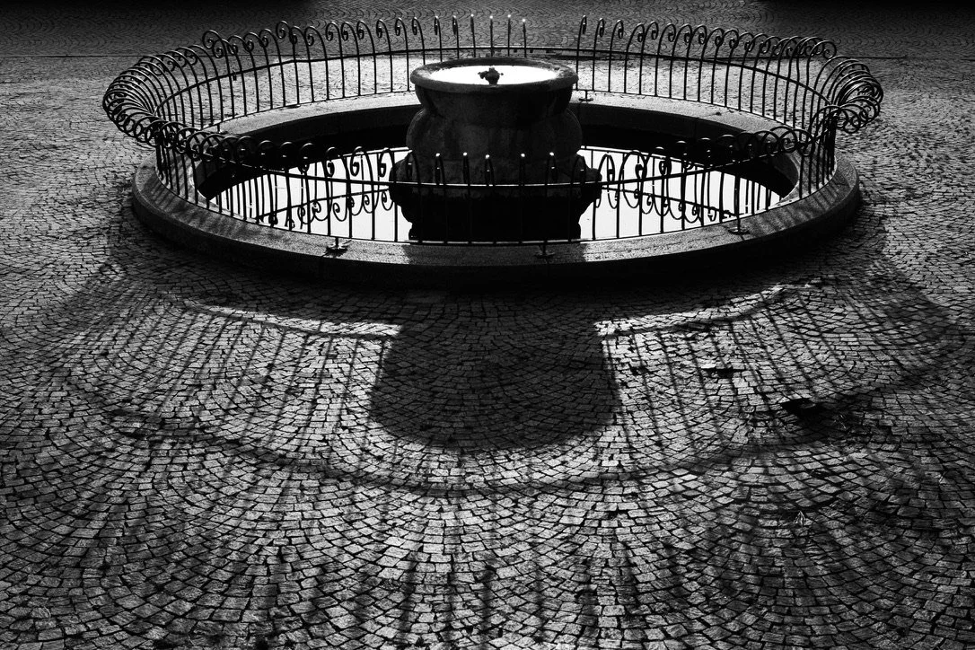 A black and white photo of a small fountain surrounded by a circular iron fence, casting a shadow on a cobblestone ground.