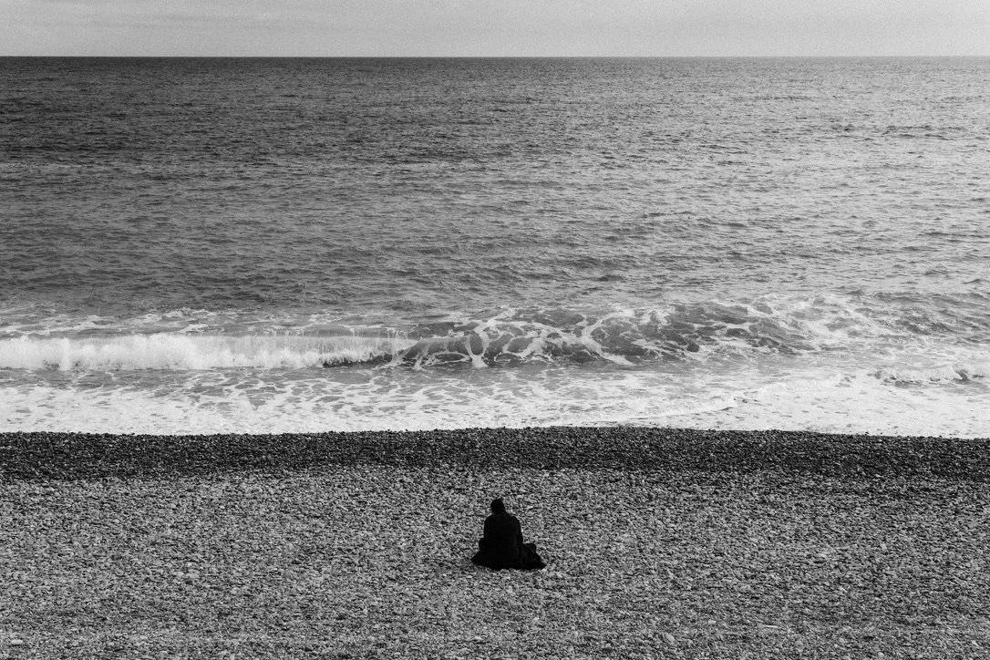 A person sitting alone on a pebble beach facing the ocean in black and white.