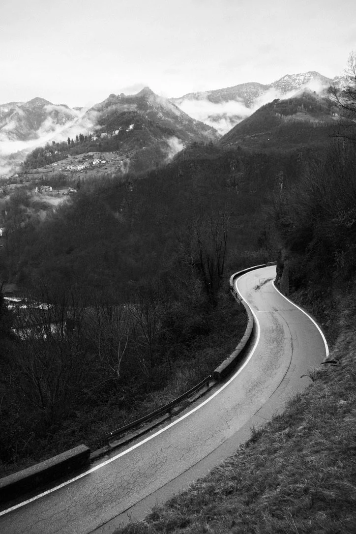 A winding mountain road with guardrails curves through a hilly landscape with trees and foggy mountain peaks in the background, in black and white.