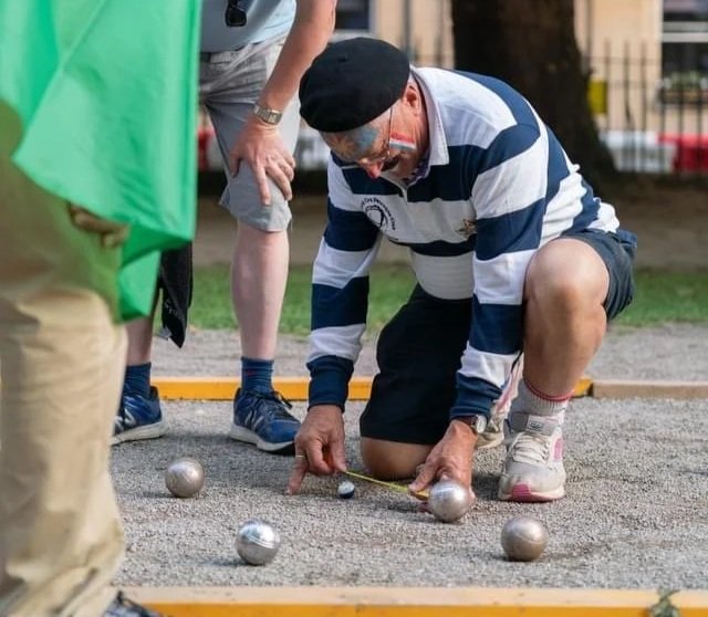 A man wearing a black beret, striped shirt, and shorts kneeling on the ground, measuring the distance between metal petanque boules during a game, with other players nearby.