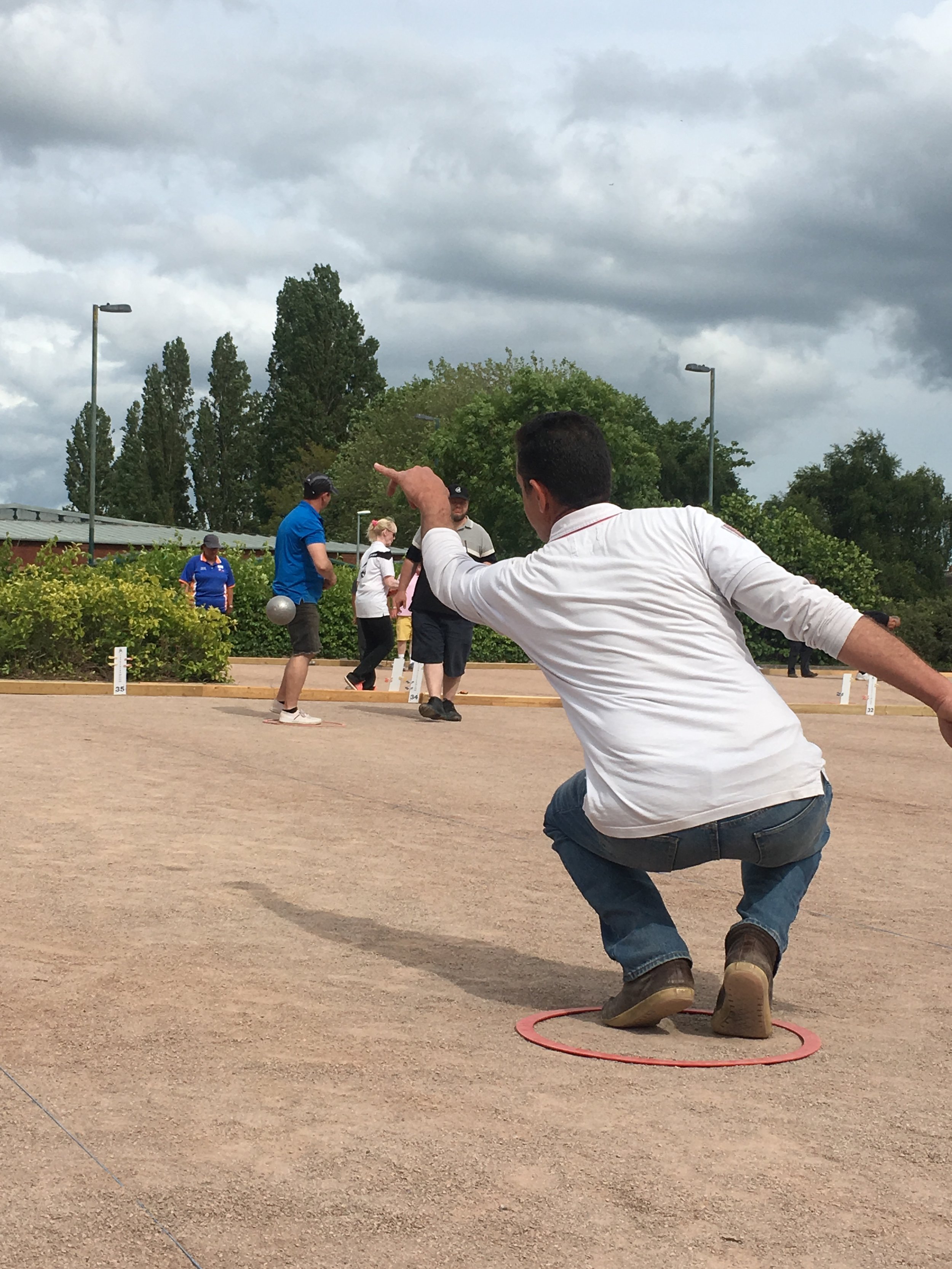 A man in a white shirt and jeans crouching on a pétanque court, pointing at the target, with several players in the background under cloudy skies.