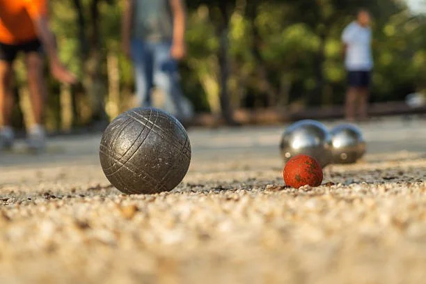 Close-up of petanque boules on a sandy court with people playing in the background.