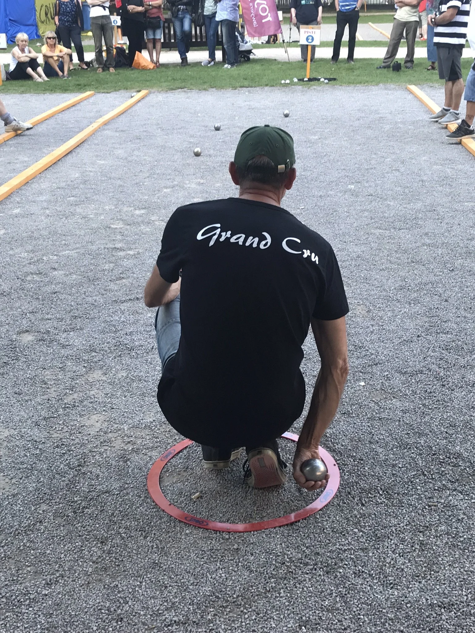 A man kneeling on the ground inside a red circle, holding a metal ball in his right hand, playing pétanque at an outdoor event with spectators watching.