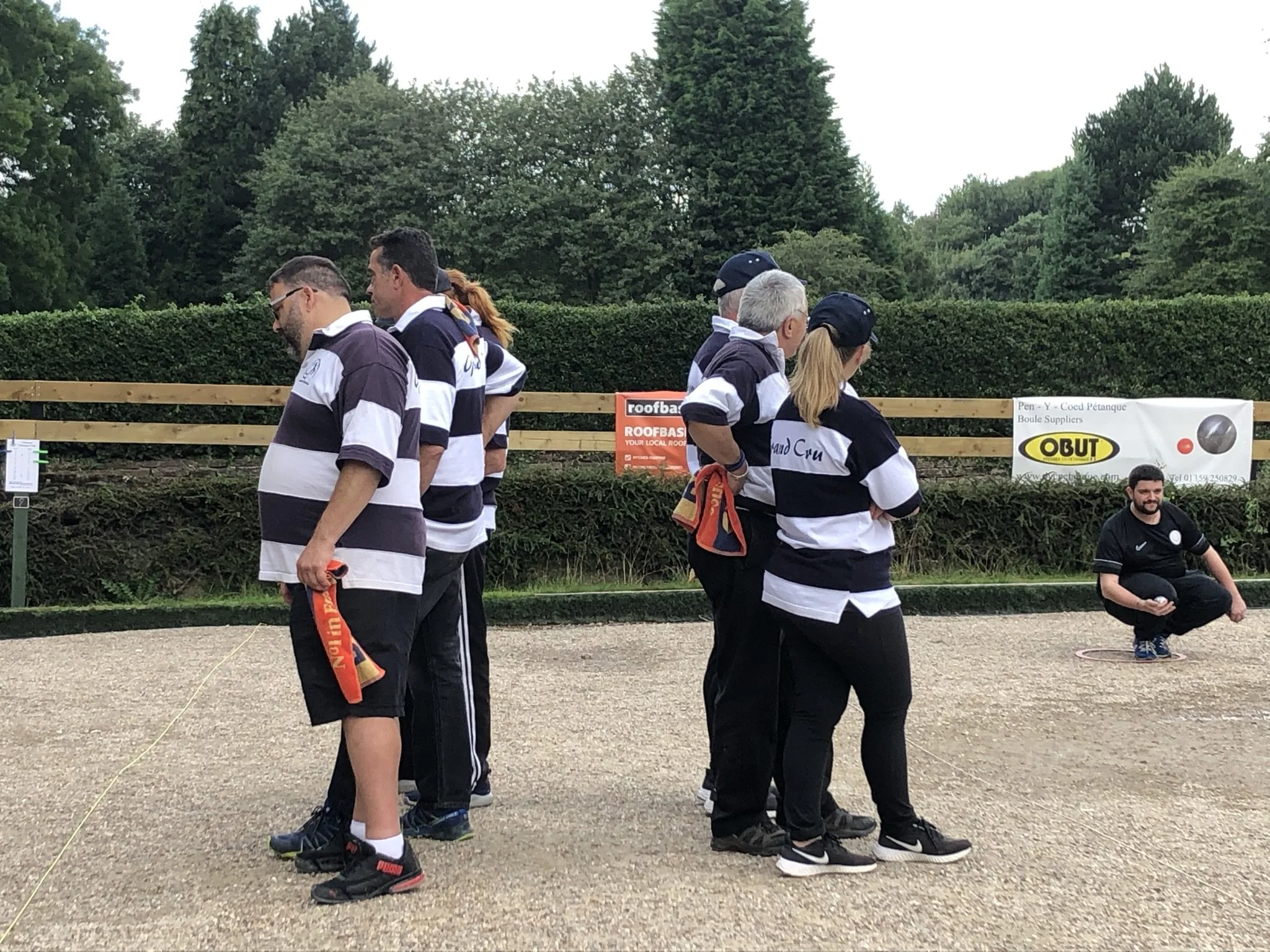 A group of seven people in black and white striped shirts gathered outdoors on a gravel court, some standing and one kneeling, with trees and advertising banners in the background.