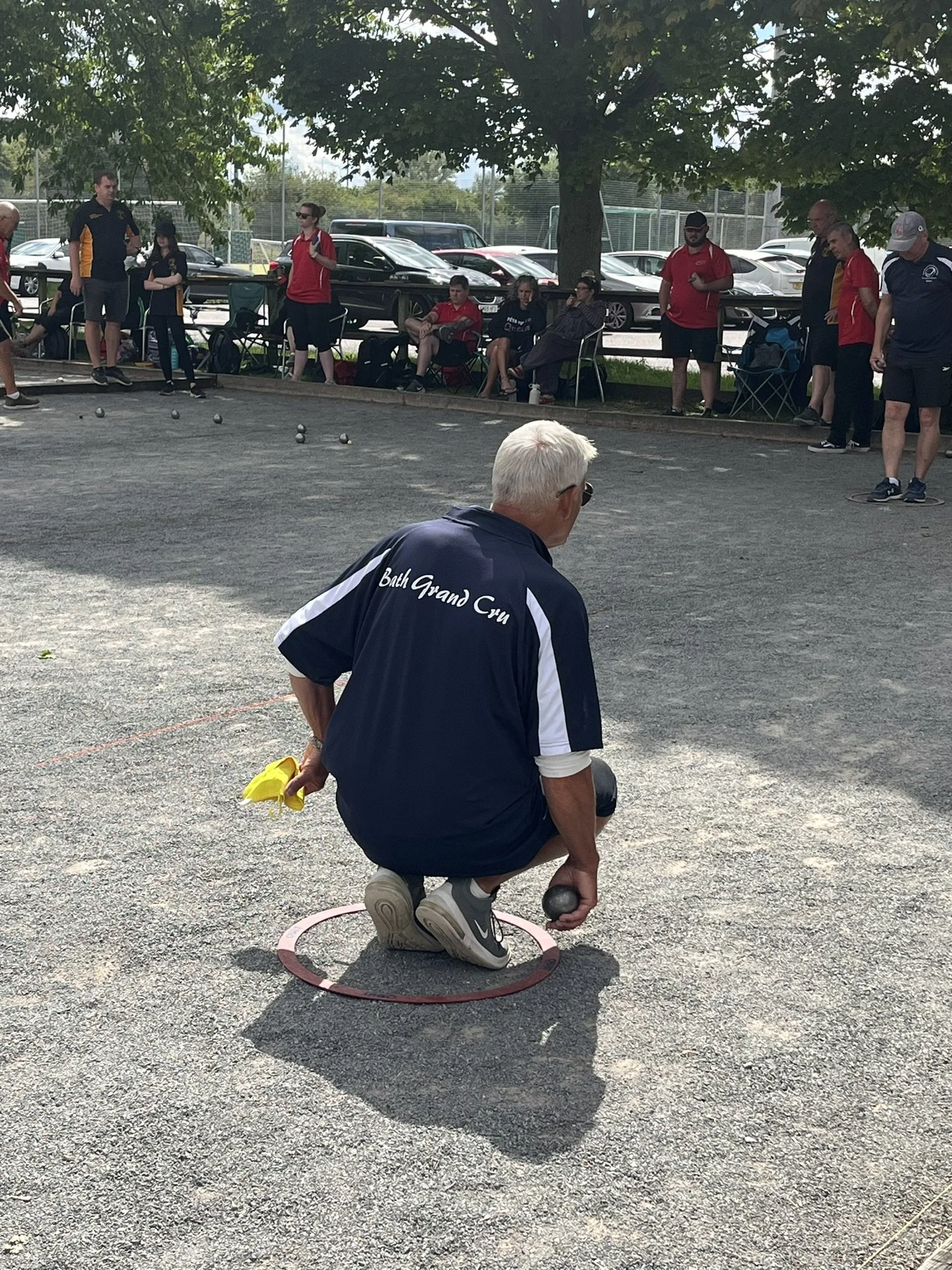 An elderly man wearing a navy blue shirt with white text on the back kneels on the ground to play pétanque, holding a metal ball in his right hand, with a yellow cloth in his left hand, and is within a red circle on the gravel surface. Behind him, several people are watching and sitting, with some standing and some seated, near a row of trees and parked cars beside a chain-link fence.
