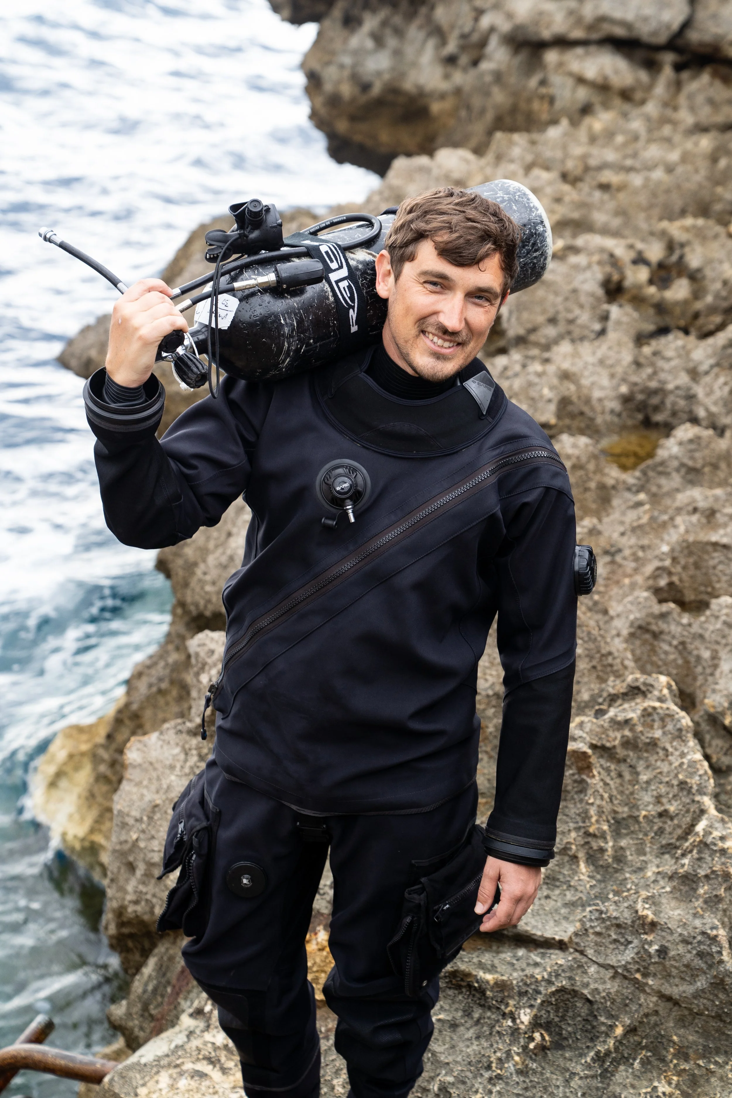 Diving instructor standing by rocks and sea holding a cylinder over his shoulder