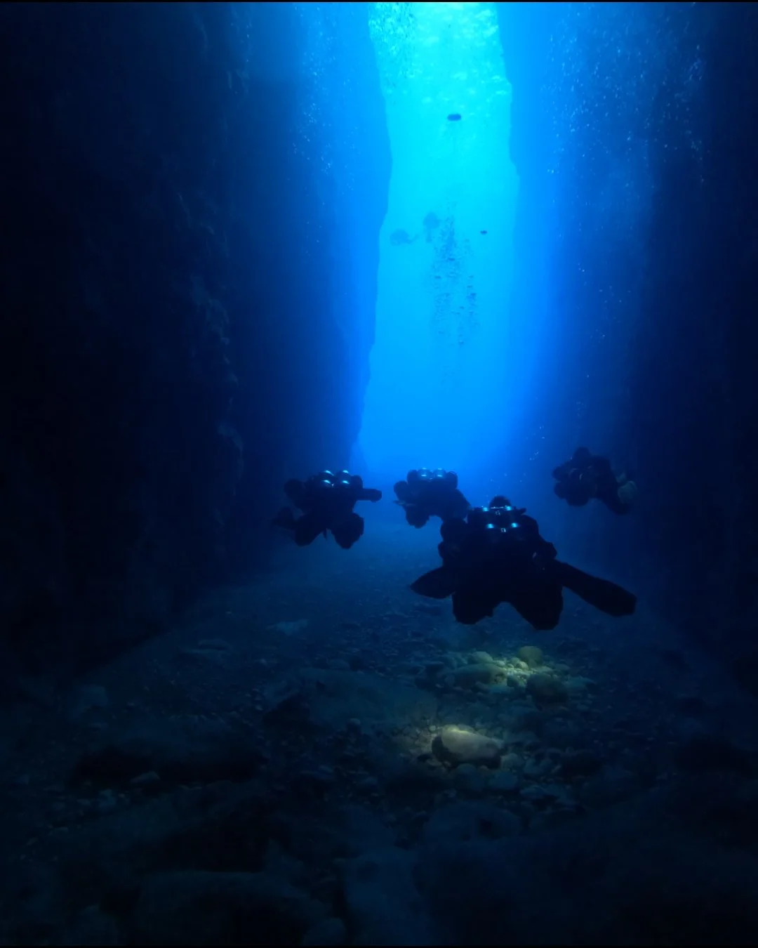 Four scuba divers swimming through an underwater canyon towards a light source.