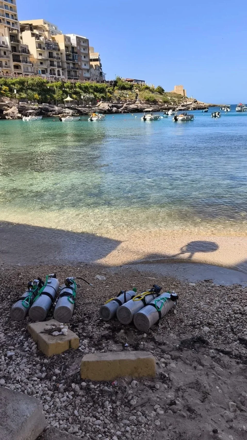 A beach scene with clear turquoise water, boats floating in the bay, and modern apartment buildings on a hillside. Several scuba tanks are resting on the sandy shore in the foreground.