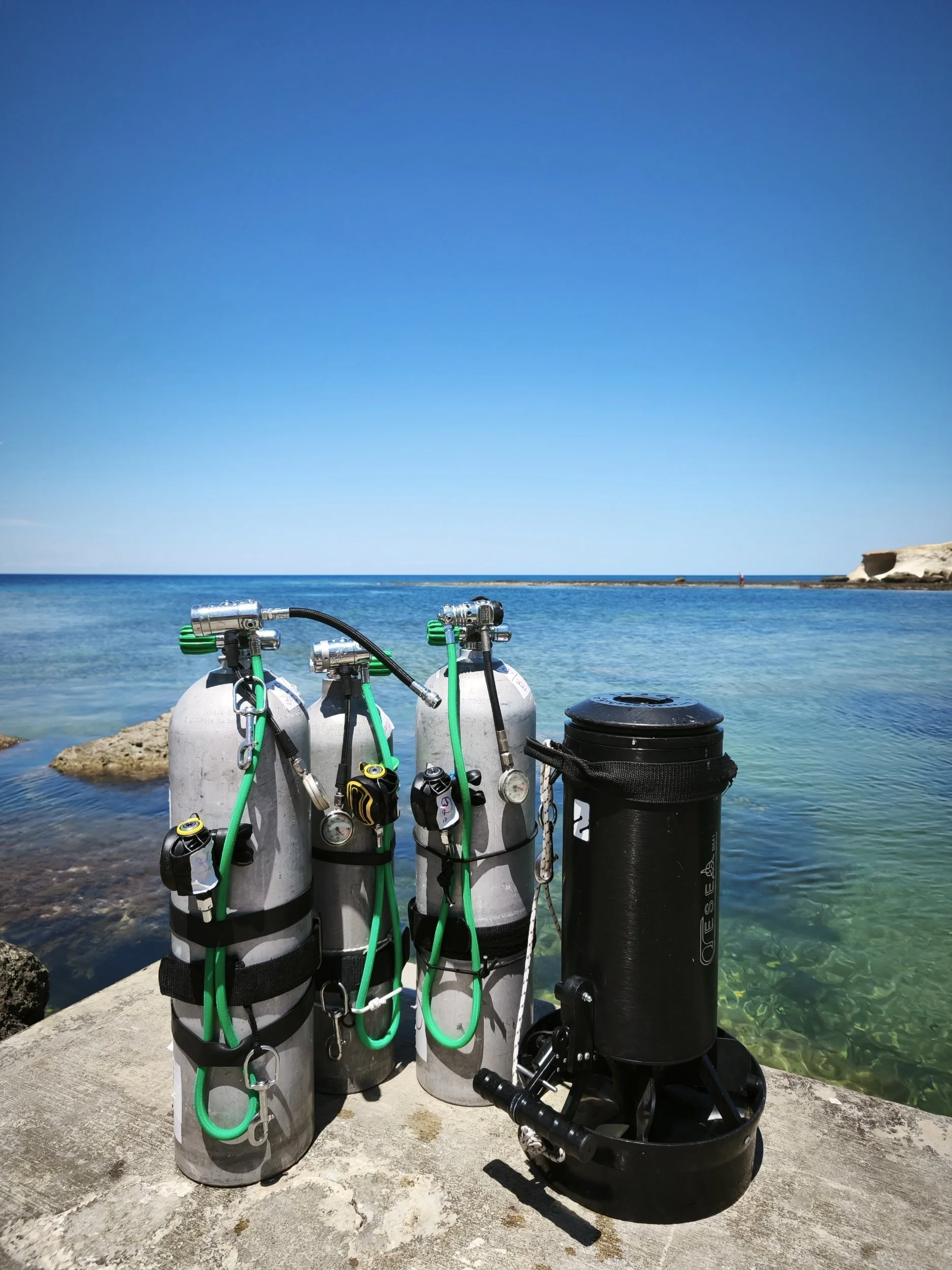 Set of scuba tanks, with regulators and hoses, on a concrete dock by clear blue ocean with a rocky shoreline and a cliff in the background under a bright sky.