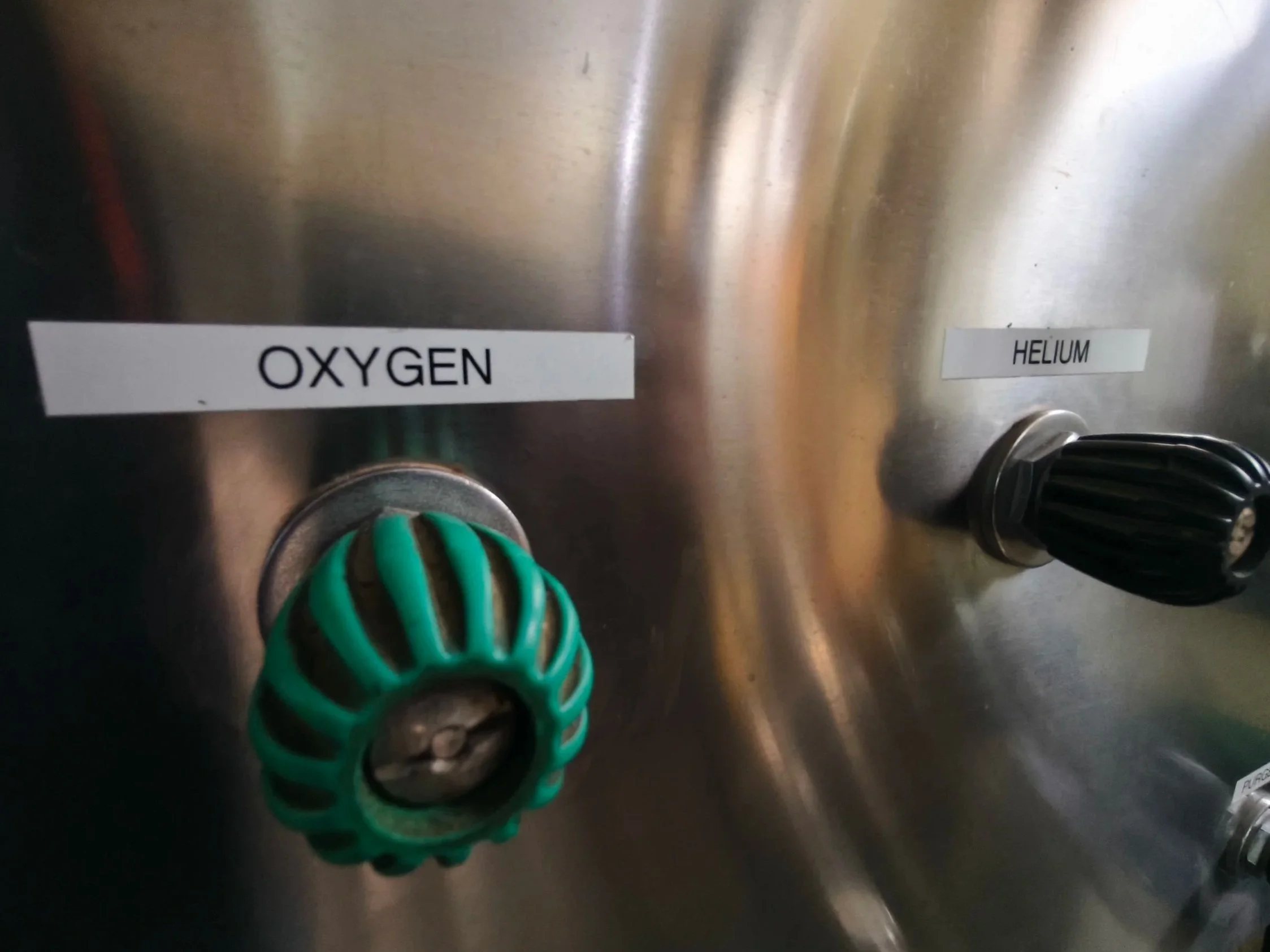 A metallic panel with two labels: 'Oxygen' and 'Helium'. A green and black valve is visible at the bottom of the panel.