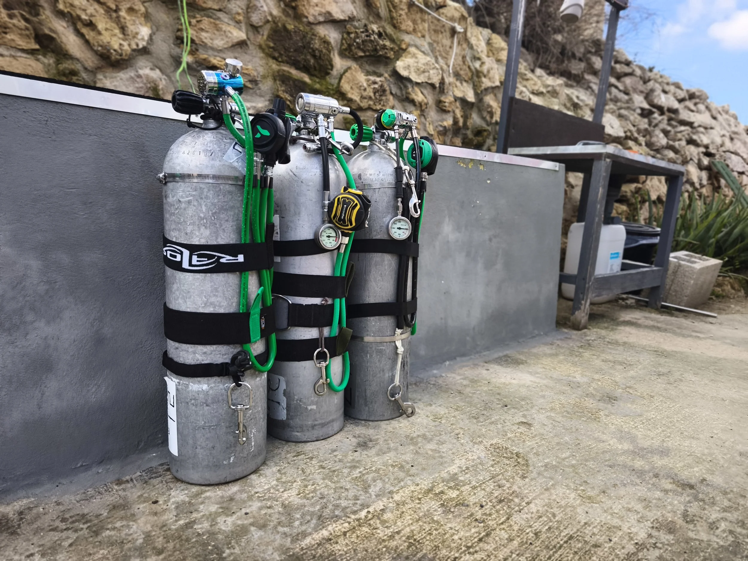 Two scuba tanks are secured with black straps and surrounded by green hoses, and are set against a concrete wall outdoors. There is a stone wall and some equipment and tools in the background.