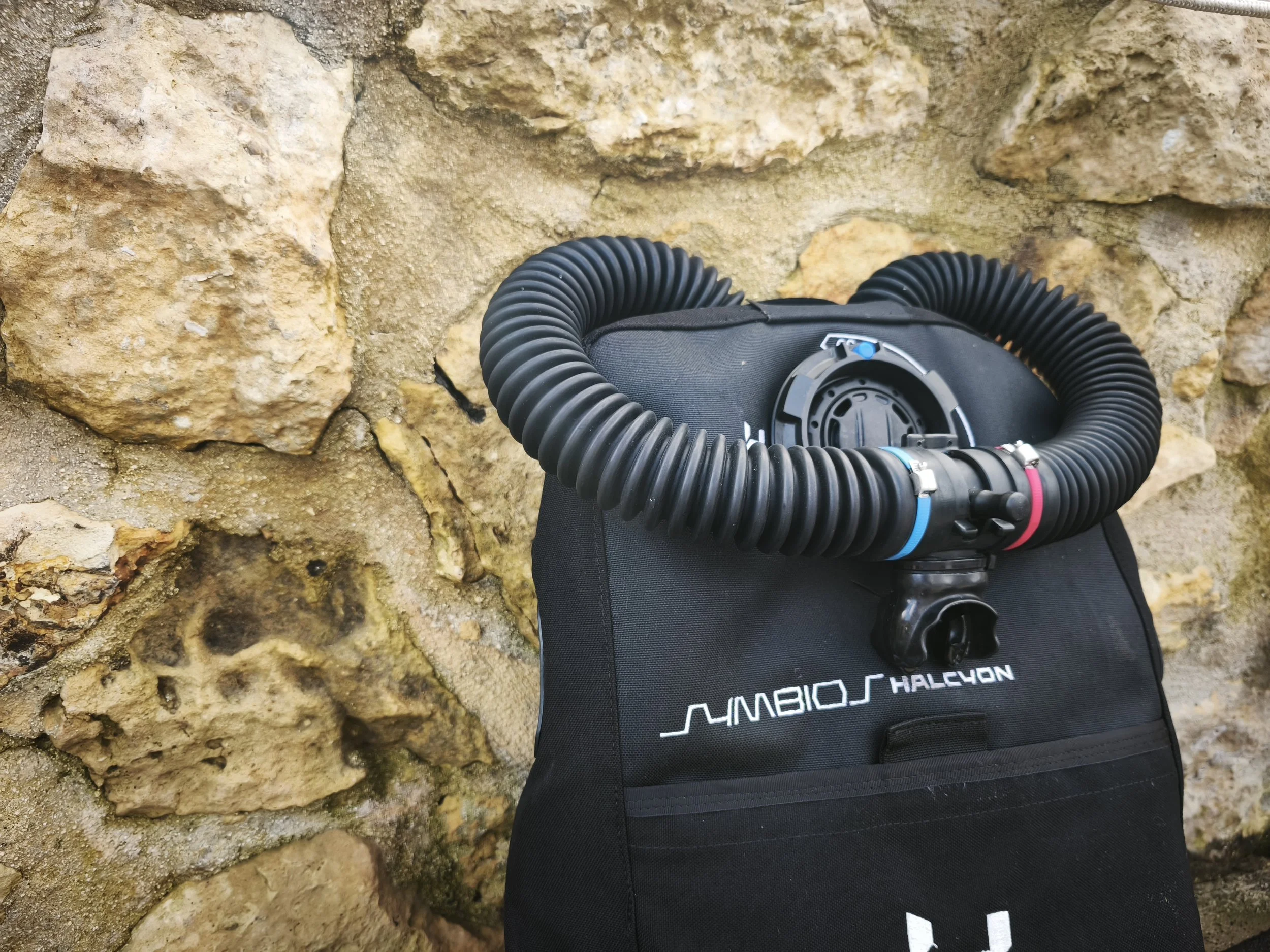 Close-up of a black diving mask with a flexible hose, resting on a black bag against a rough, beige stone wall.