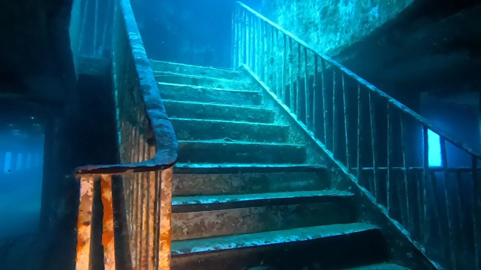 Underwater stairs with steel handrails, rusted and covered in algae, leading upward in a submerged structure with blue lighting.