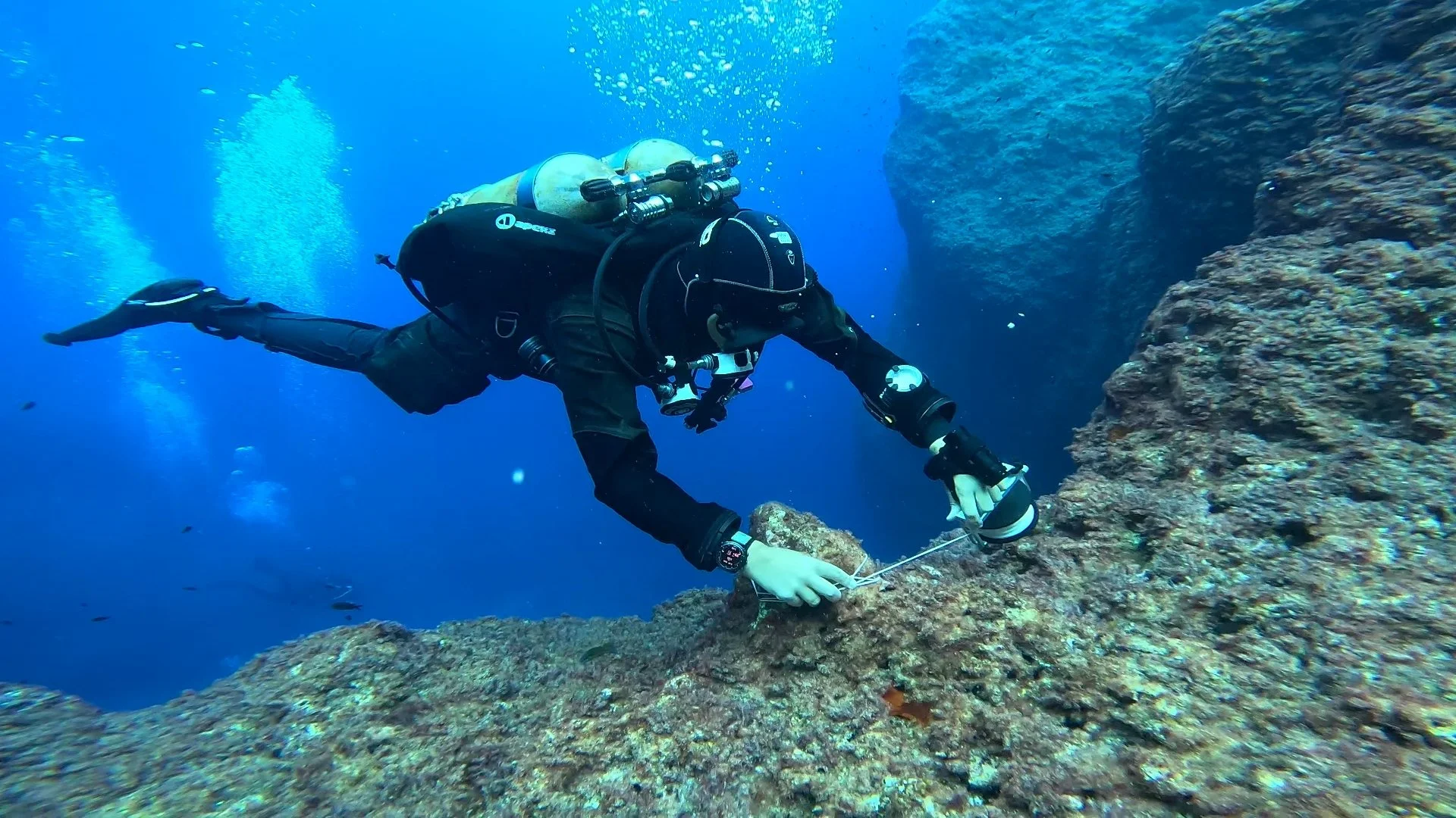 A scuba diver in black gear exploring underwater near rocks.