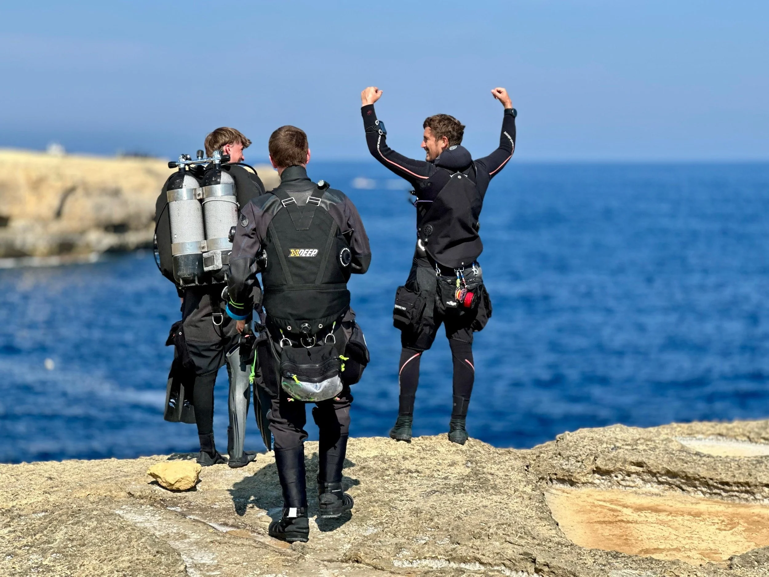 Three scuba divers standing on a rocky beach near the ocean, two facing away from the camera with gear on and one facing them with arms raised in celebration.