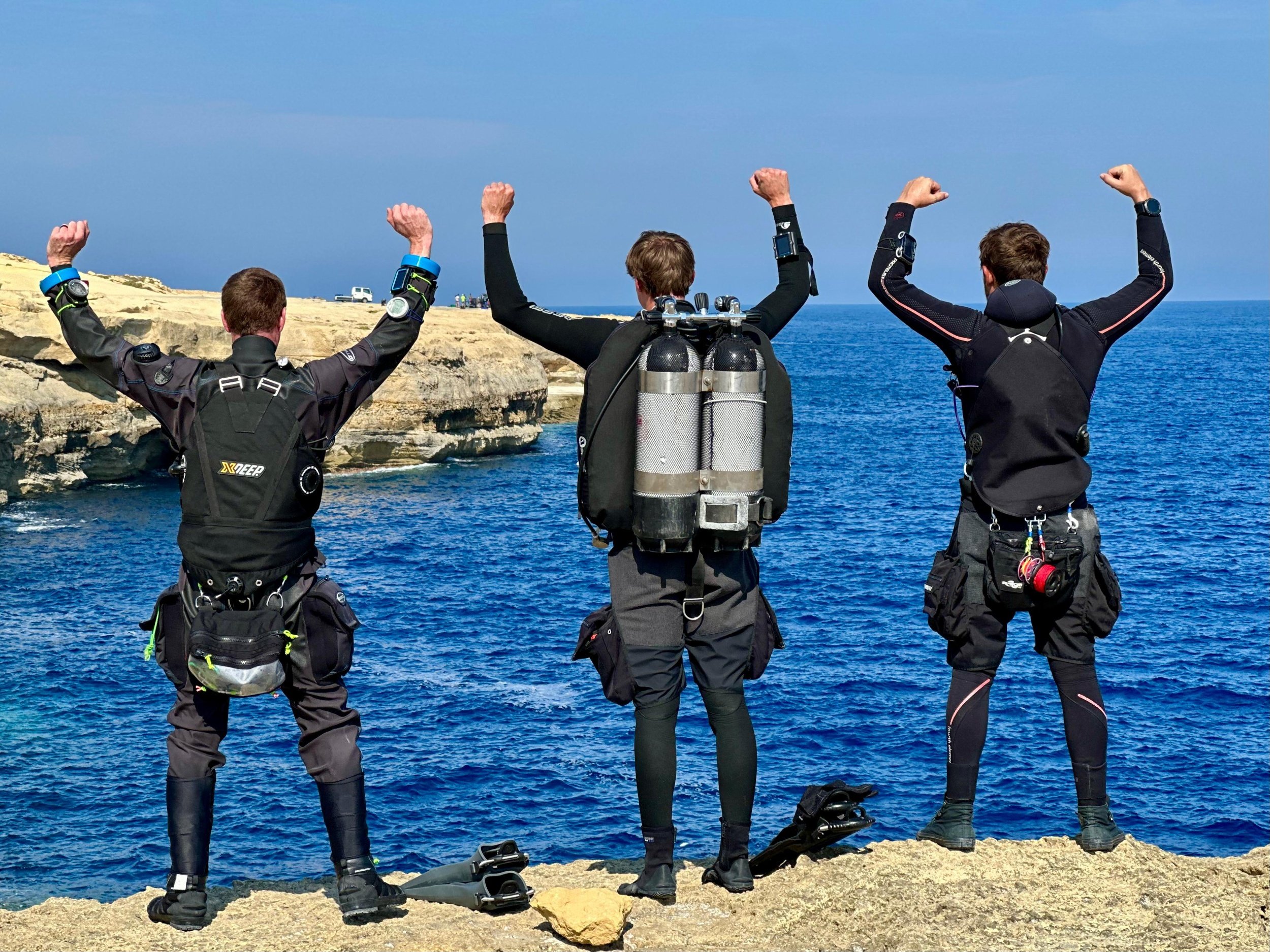 Three scuba divers in black suits and fins standing on rocks by the ocean, with coastal cliffs and vehicles in the background.