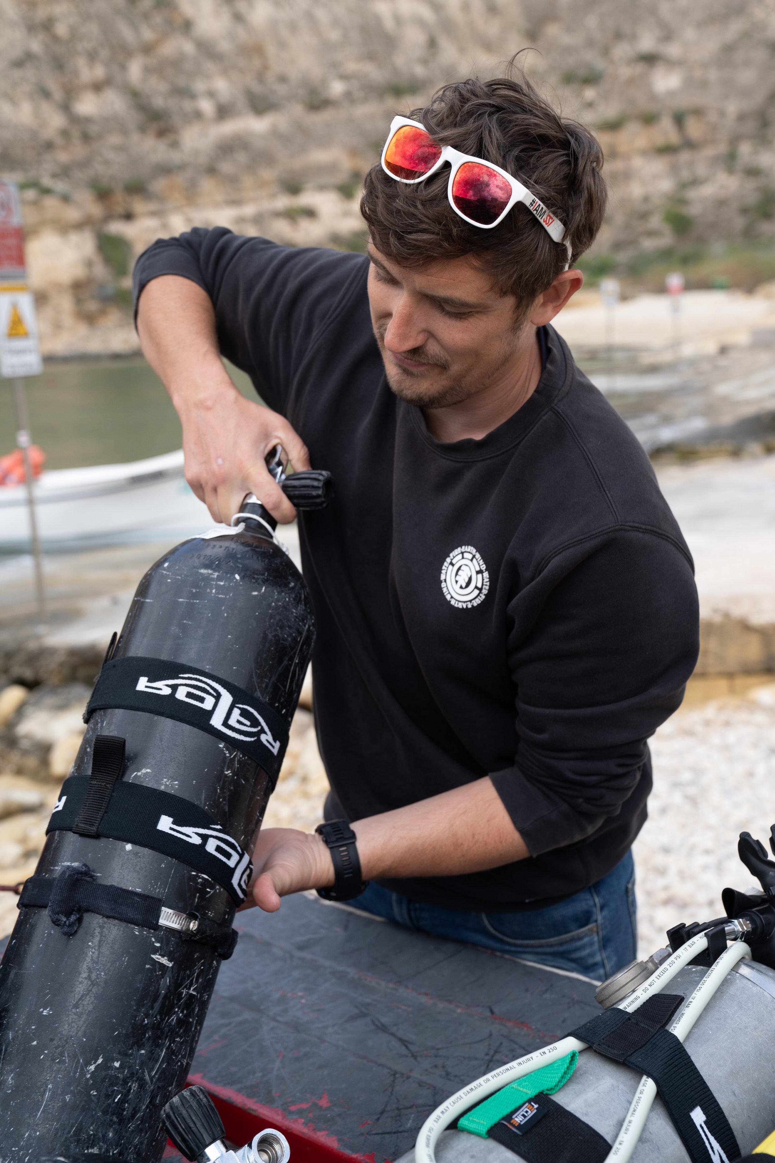 A man adjusting scuba diving gear on a boat with a rocky hillside in the background.