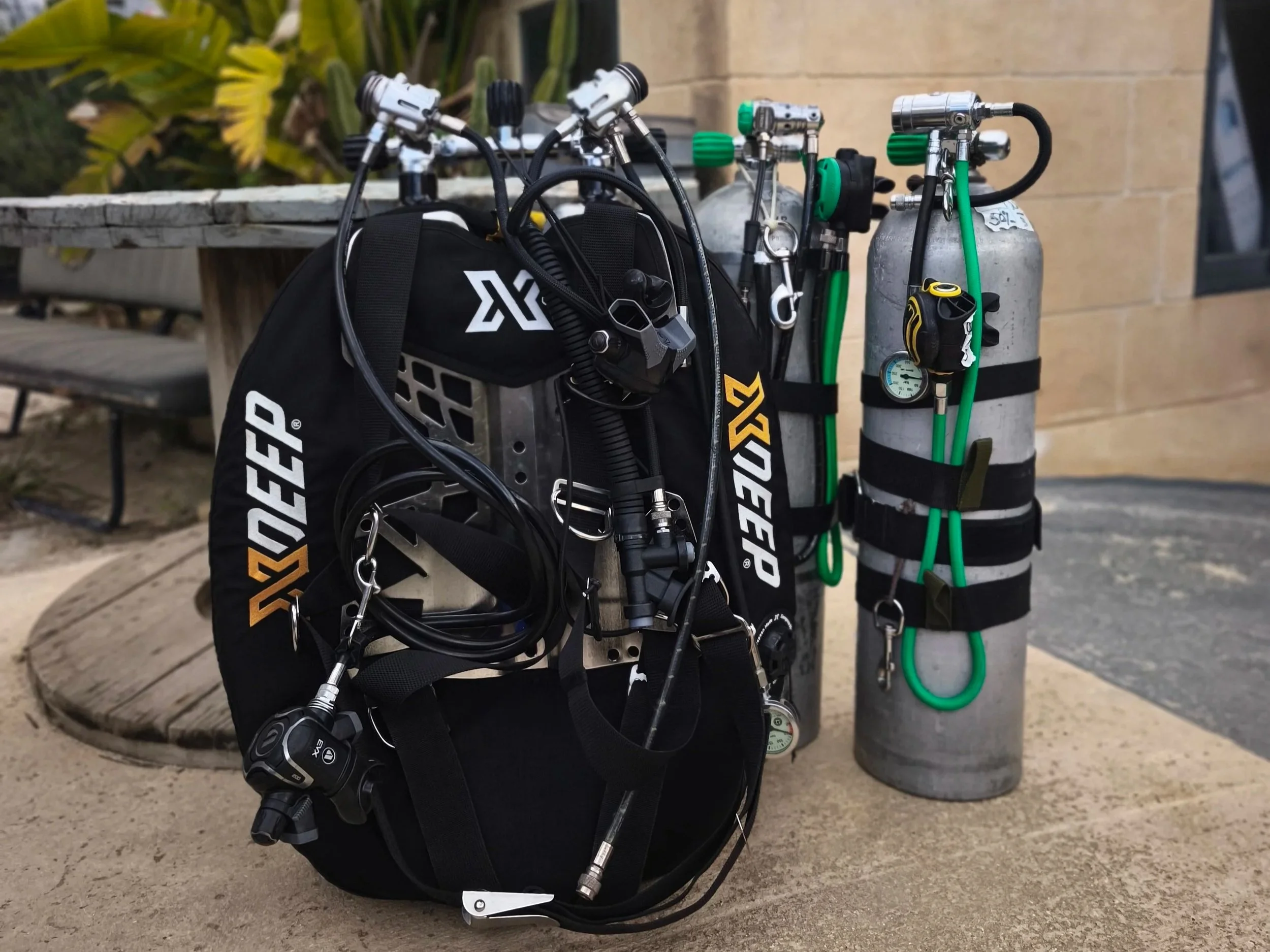 Scuba diving gear including tanks, regulator, and buoyancy control device, stored outside near a wooden bench and a building wall.