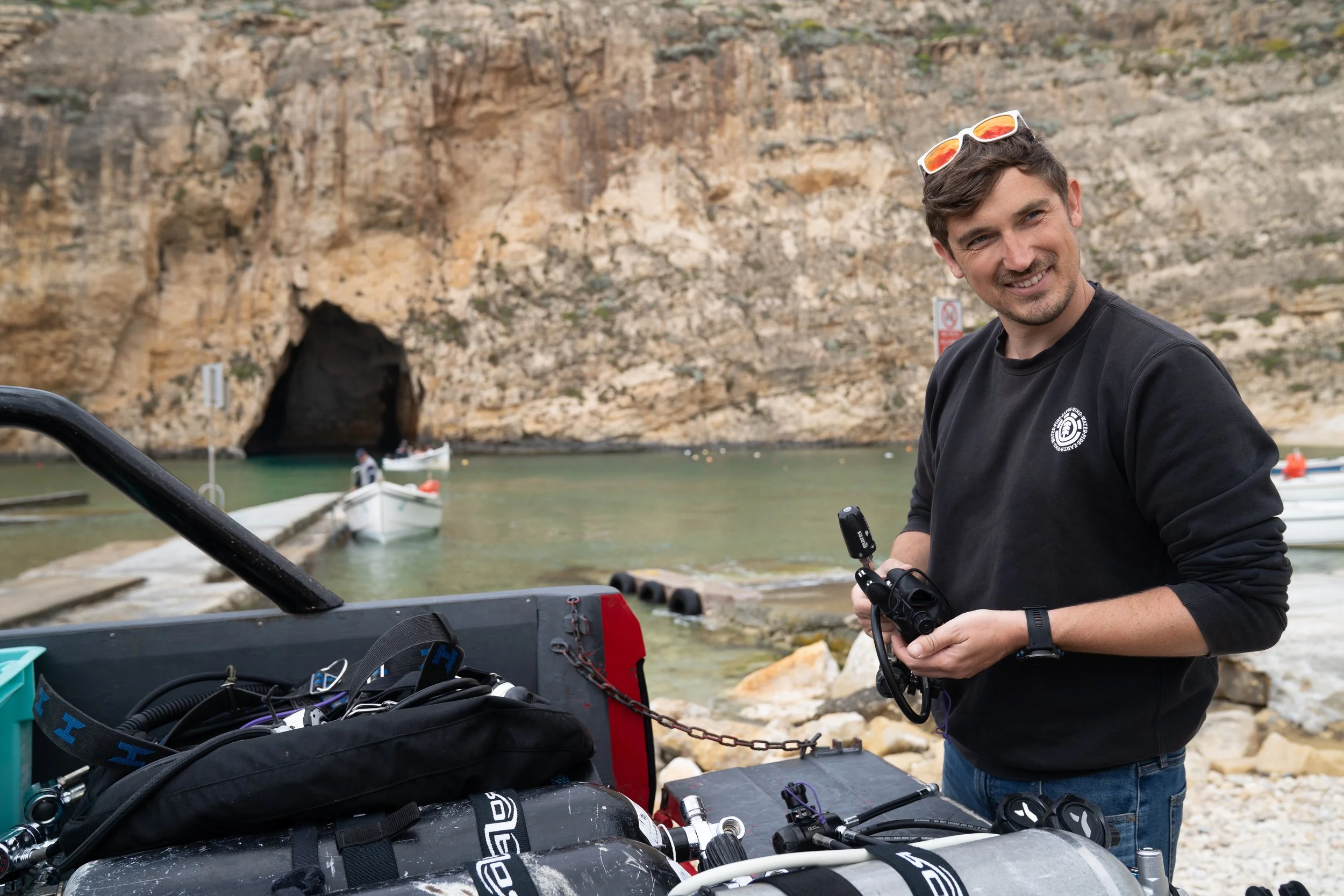 A young man with sunglasses on his head standing near boats and diving equipment by a rocky coastline with a cave entrance and water in the background.