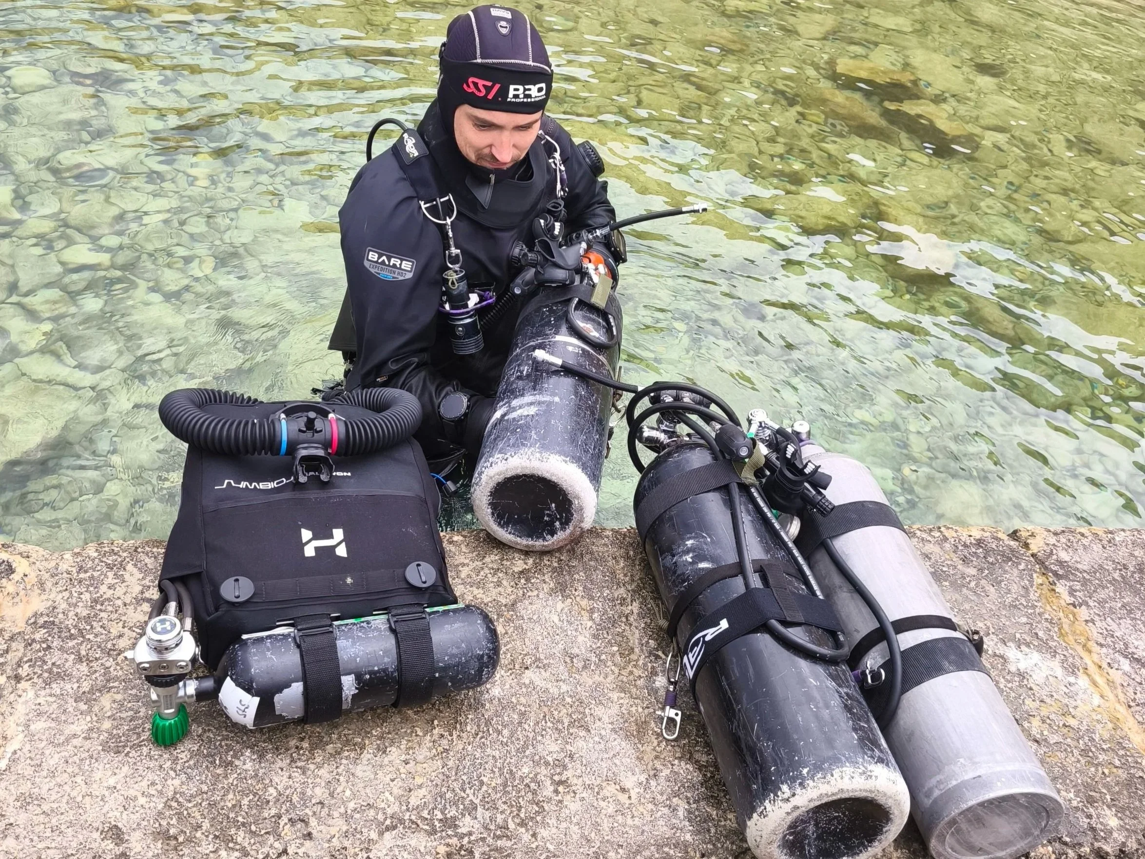 A diver in a black wetsuit and beanie hat with the logo 'SSI PADI' on it, sitting in shallow clear water with a rocky bottom, surrounded by scuba diving equipment including tanks and regulator hoses.