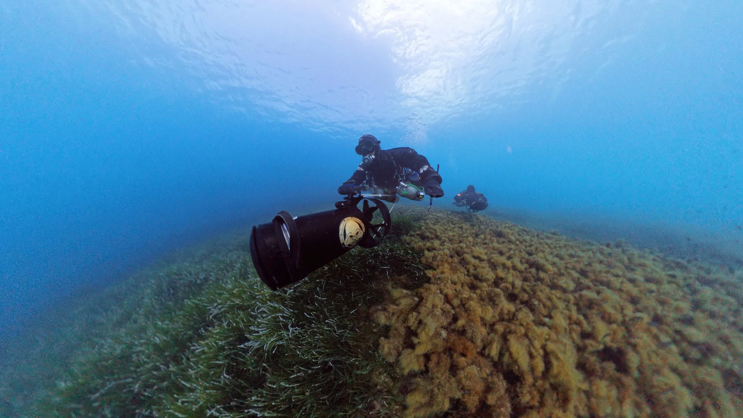 Two scuba divers exploring an underwater coral reef. One diver is in the foreground holding a camera or flashlight, while the second diver is further back. The water above is clear with sunlight filtering through, illuminating the scene.