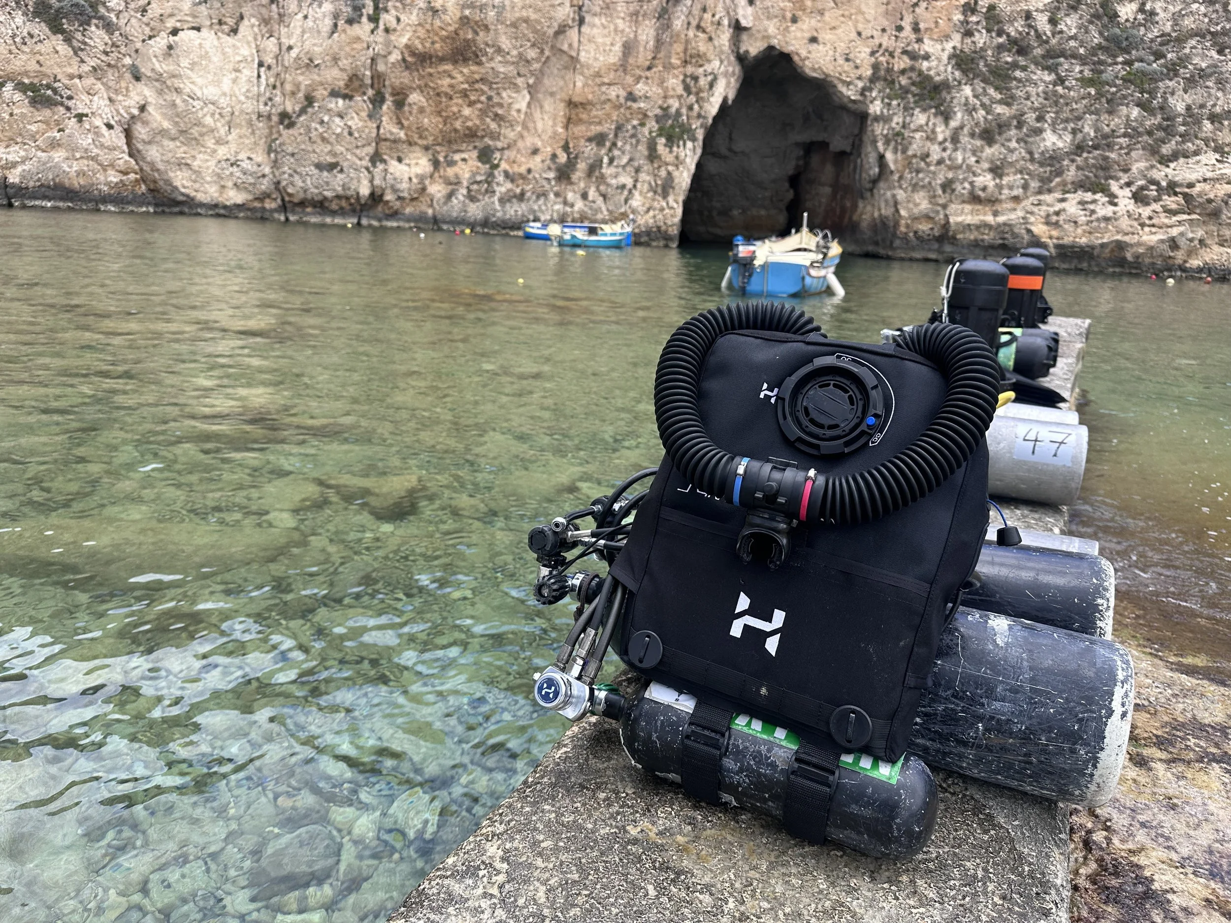 View of a small boat docked near a rocky cave entrance with clear water and rocks along the shoreline, and scuba diving gear in the foreground.