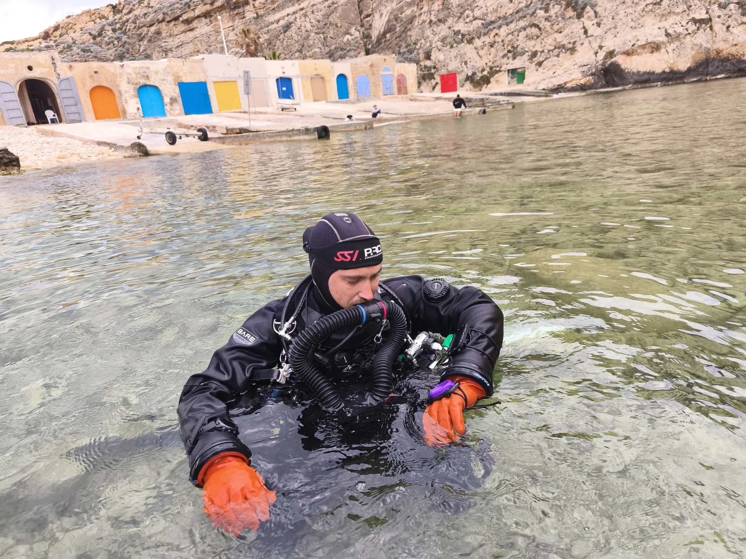 A man in a wetsuit and diving gear in clear water, with a colorful building and rocky hillside in the background.