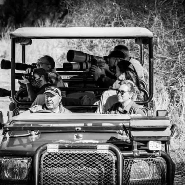 Group of people riding in a safari vehicle, with some using binoculars or cameras, outdoors near trees.