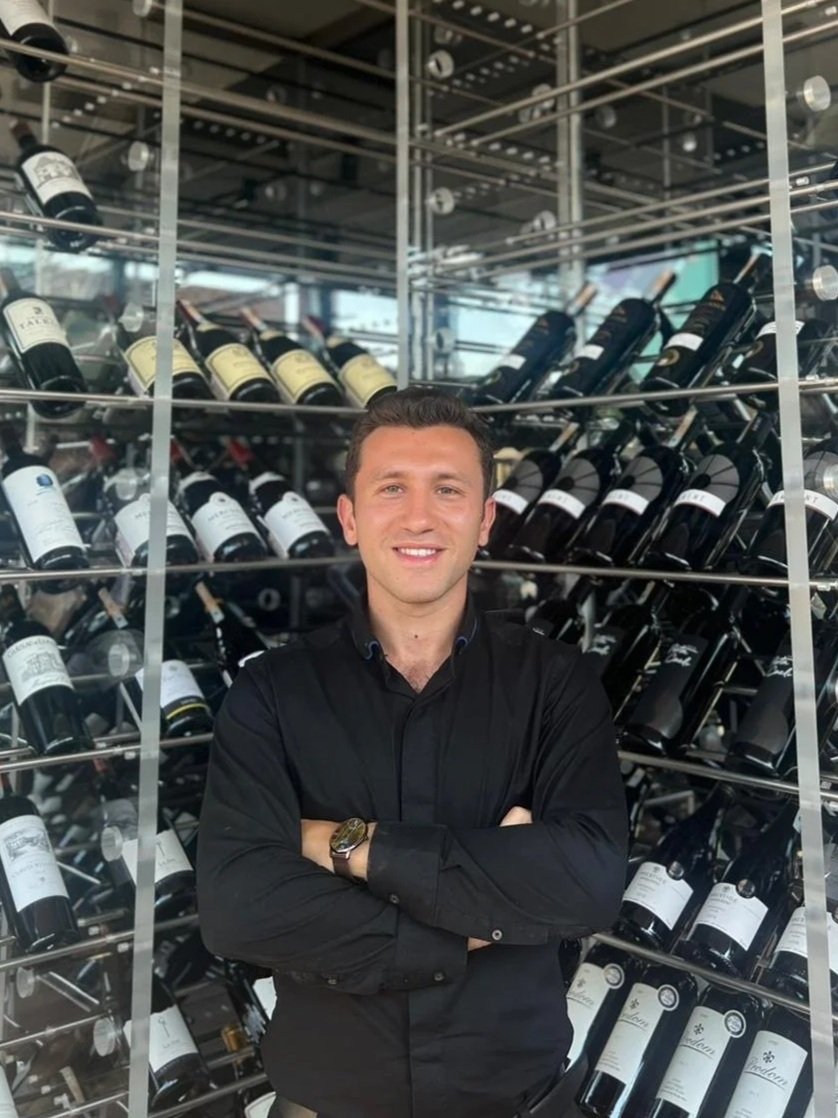A man in a black shirt standing with arms crossed in front of a metal wine rack filled with bottles of wine.