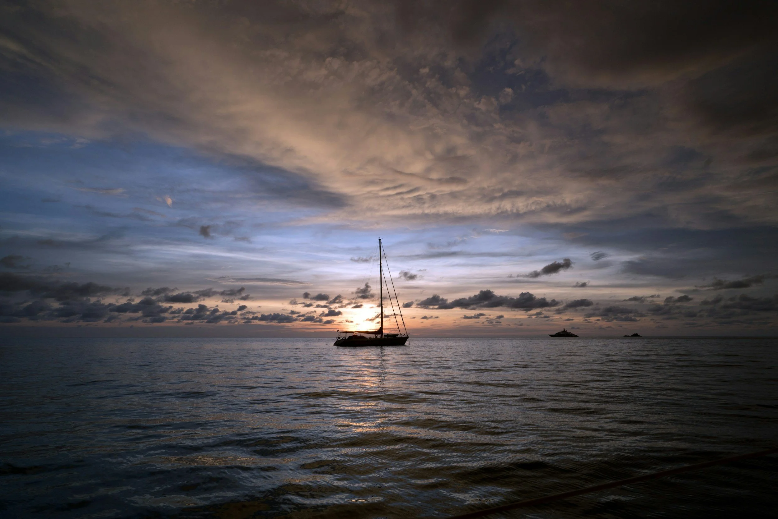A sailboat floating on calm ocean waters during sunset with a dramatic sky filled with clouds.